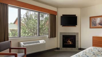 Ein Hotelzimmer mit Kamin, einem großen Fenster mit Vorhängen, einem Bett, einem Sessel und einem flachbildfernseher an der Wand.