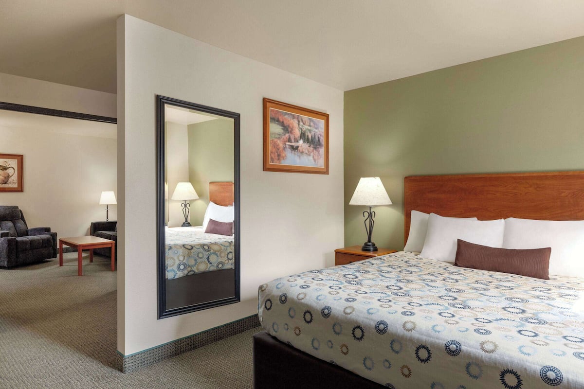 A hotel room with a bed, a sitting area with a chair and table, a nightstand with a lamp, and a painting on the wall.