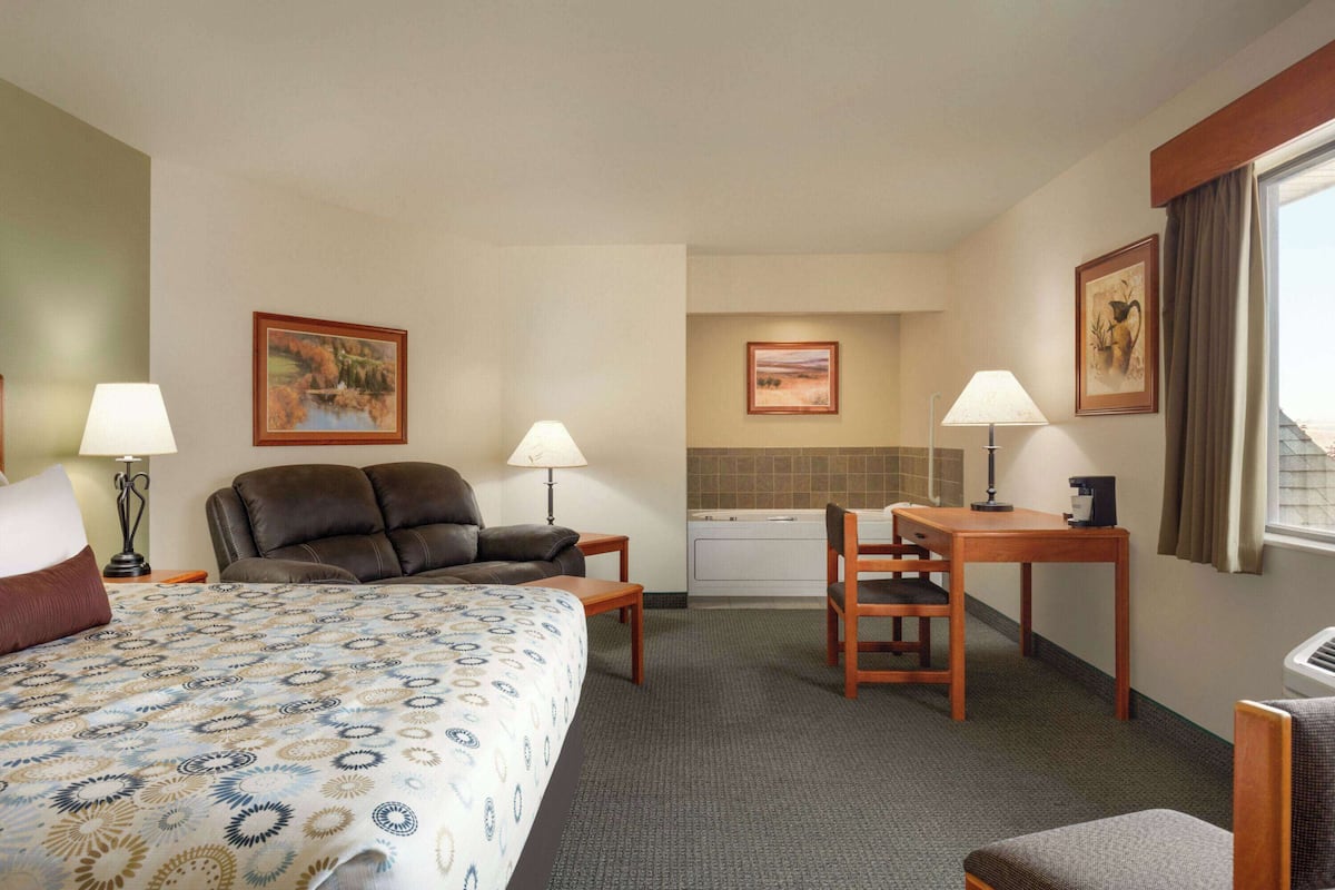 A hotel room with a bed, two chairs, a small table, a desk, a window with curtains, and a kitchenette in the background.