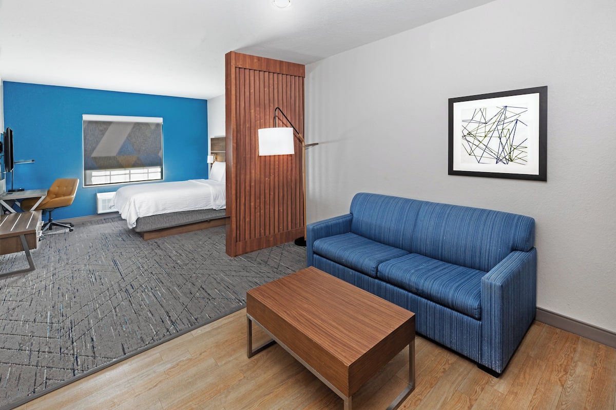 A modern hotel room with a blue accent wall, a wooden partition, a blue sofa, a wooden coffee table, a bed with white bedding, a desk with a chair, and a framed abstract artwork on the wall.