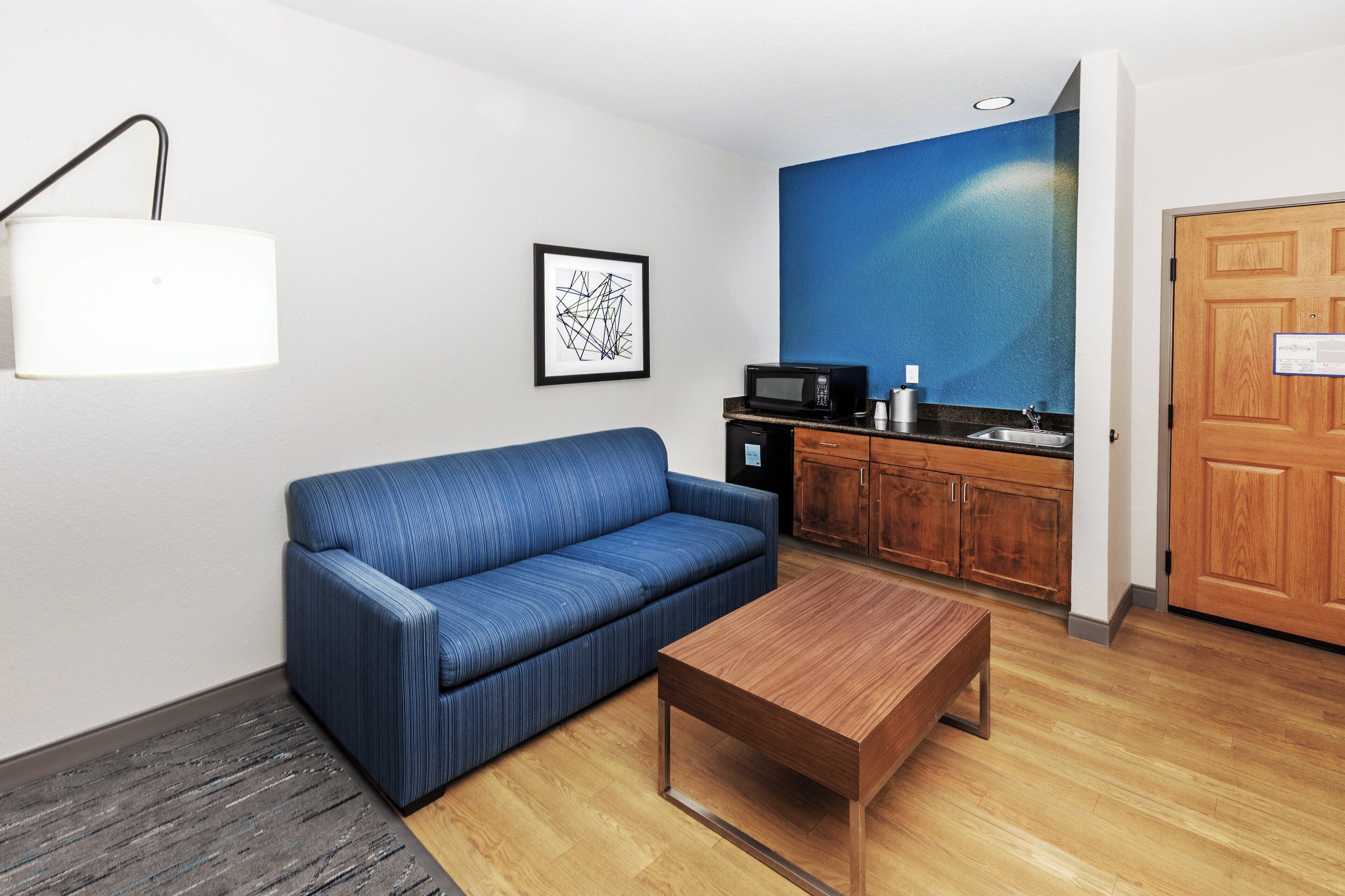 A room with a blue sofa, a wooden coffee table, a microwave, and a kitchen area with wooden cabinets.