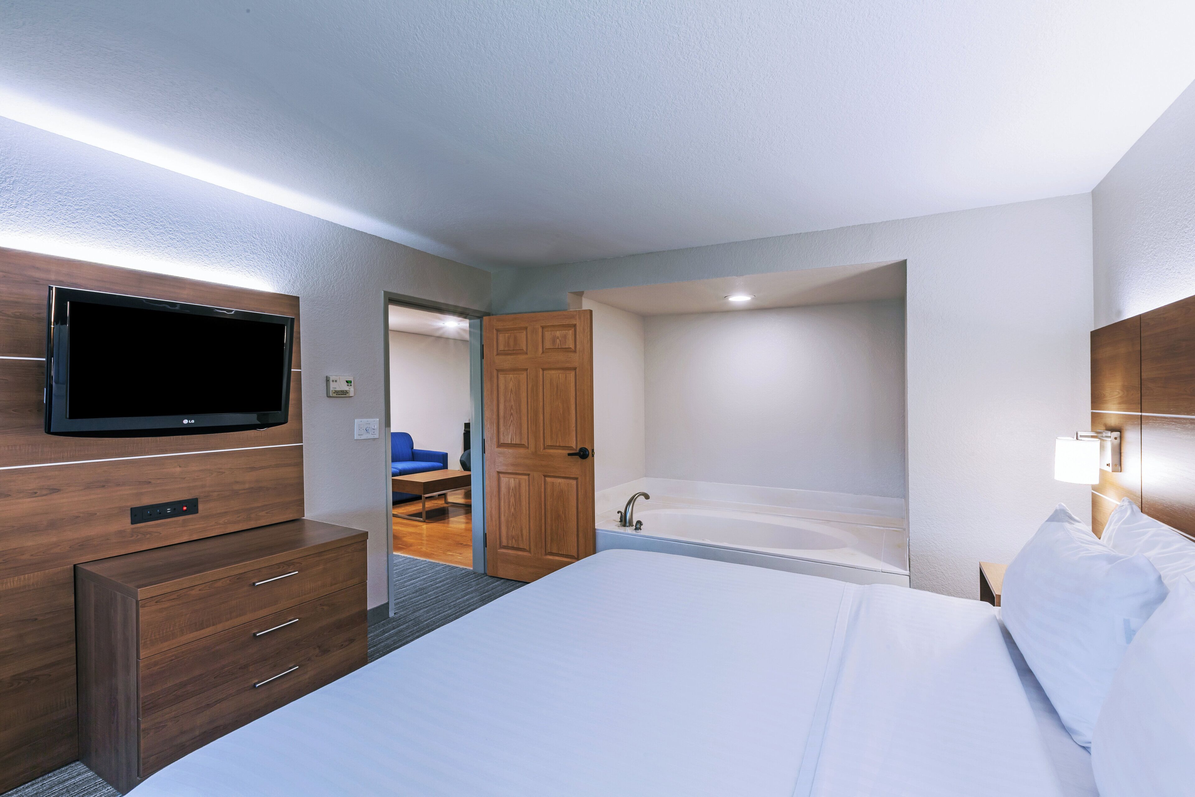 Ein Hotelzimmer mit einem Flachbildfernseher, einer hölzernen Kommode, einem Bett mit weißer Bettwäsche, einem Badezimmer mit Badewanne und einem blauen Sofa im angrenzenden Raum.