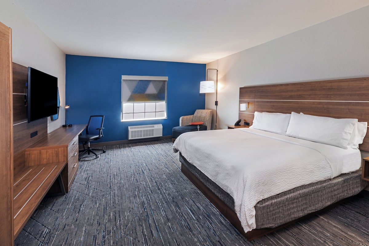 A hotel room with a large bed, a desk with a chair, a television, and a window with blinds.