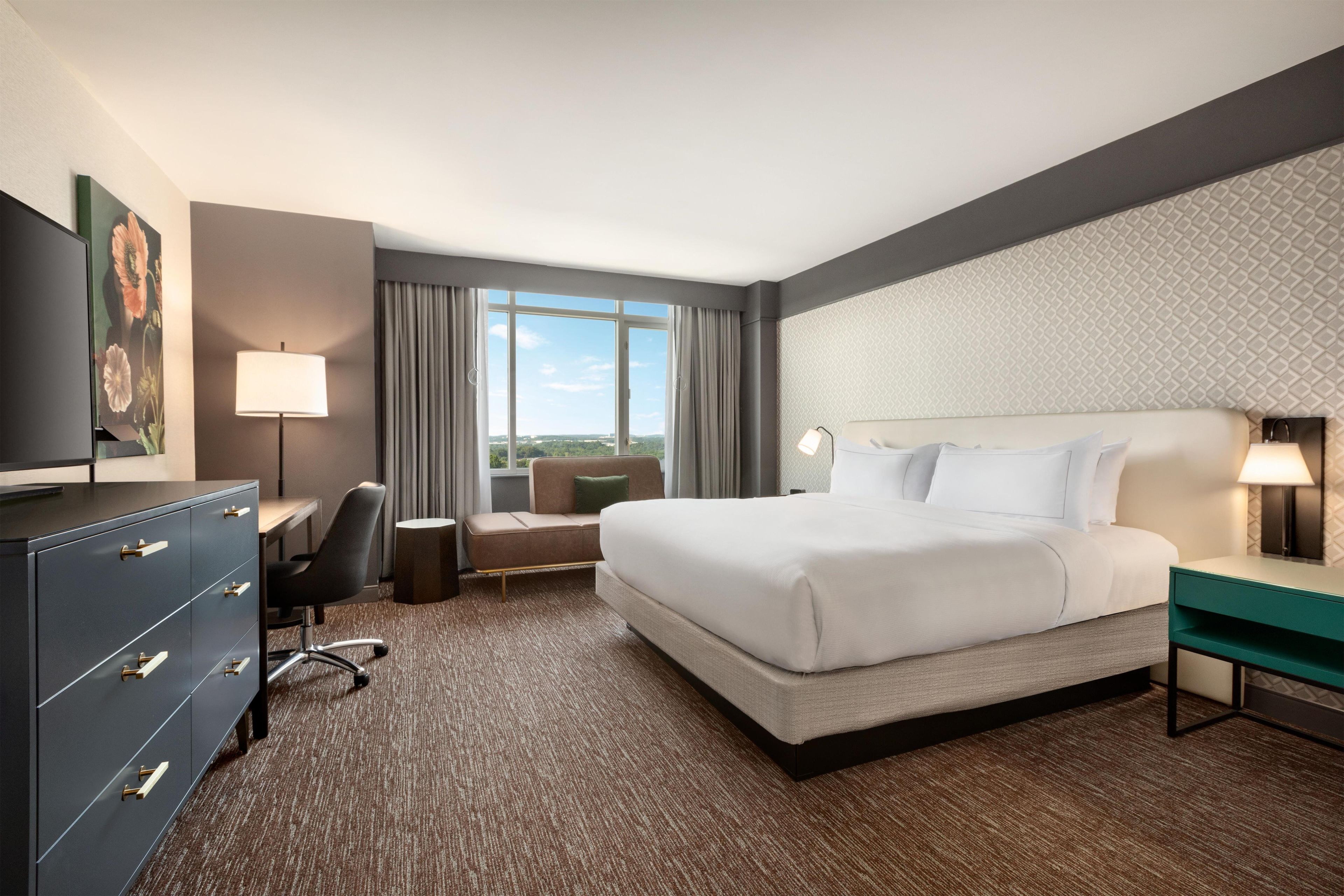A hotel room with a large bed, a desk, a chair, a dresser, and a view of the outdoors.