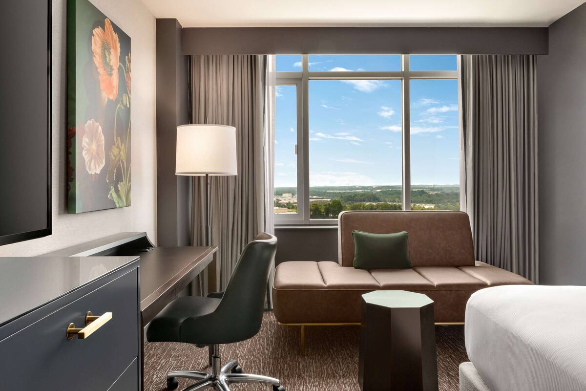 A hotel room with a bed, a desk with a chair, a sofa, a large window with a view, and a painting on the wall.