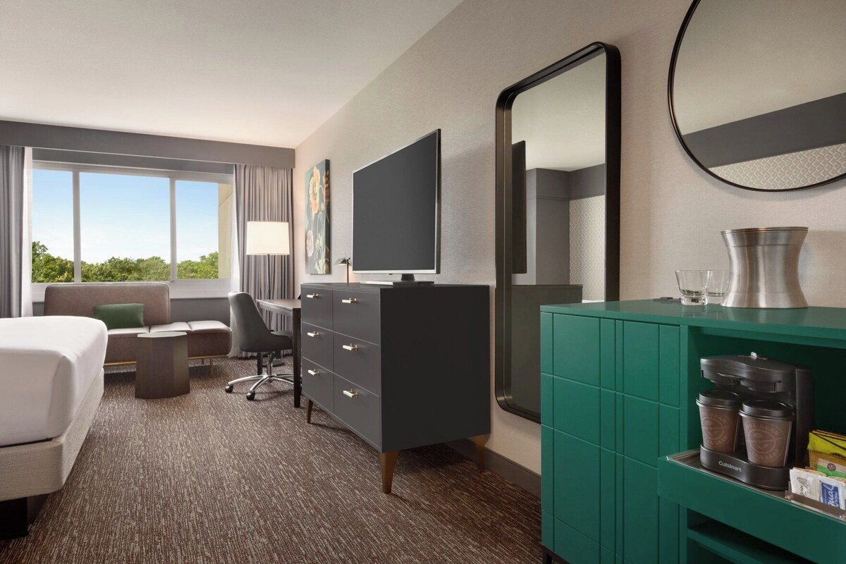 A hotel room with a bed, desk, chair, dresser, television, mirror, and a kitchenette area with a coffee maker and storage containers.