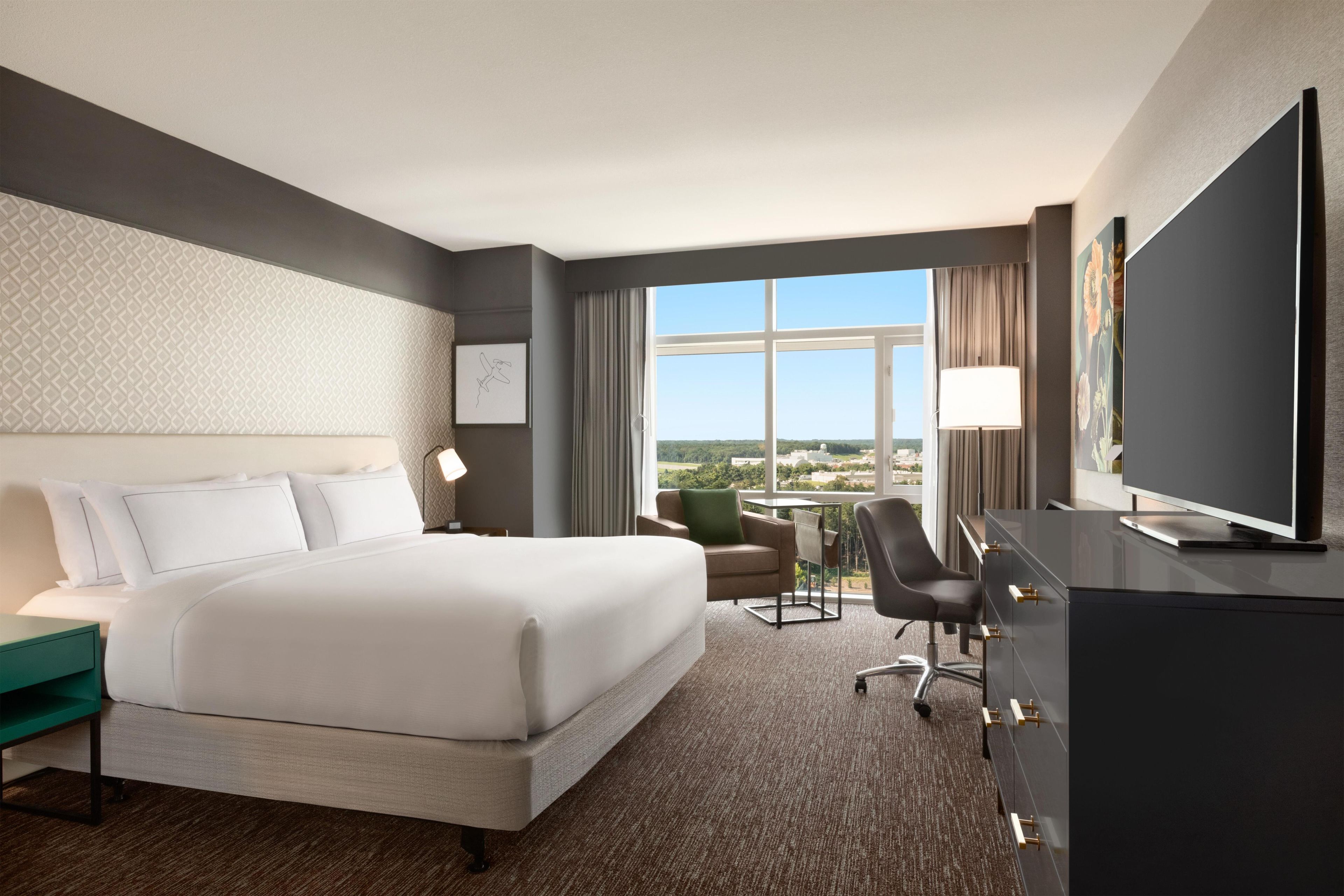 Ein Hotelzimmer mit einem großen Bett, einem Schreibtisch mit Flachbildfernseher, einem Sessel, einem kleinen Tisch und Blick auf die Stadt.