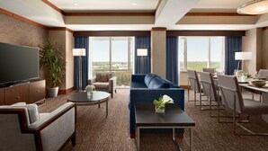 A hotel room with a large flat-screen TV, a sofa, a dining table with chairs, a potted plant, and large windows with curtains. - Hilton Baltimore BWI Airport (Linthicum Heights)