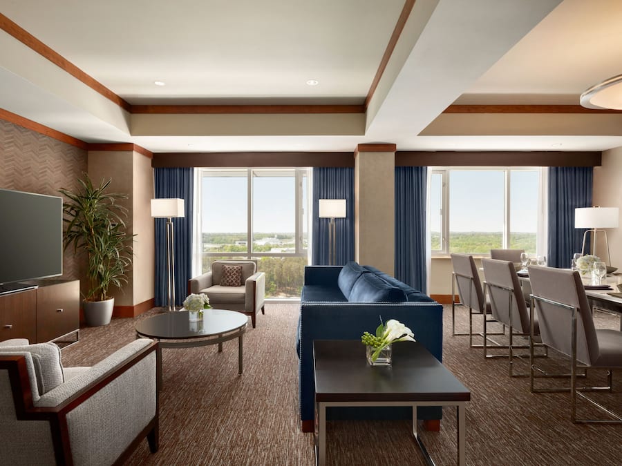 A hotel room with a large flat-screen TV, a sofa, a dining table with chairs, a potted plant, and large windows with curtains.