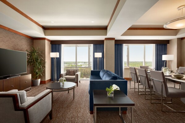 A hotel room with a large flat-screen TV, a sofa, a dining table with chairs, a potted plant, and large windows with curtains. - Hilton Baltimore BWI Airport (Linthicum Heights)