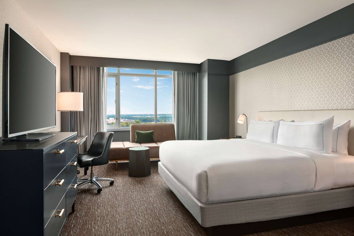 A hotel room with a large bed, a TV, a desk with a chair, and a view of the city through the window.