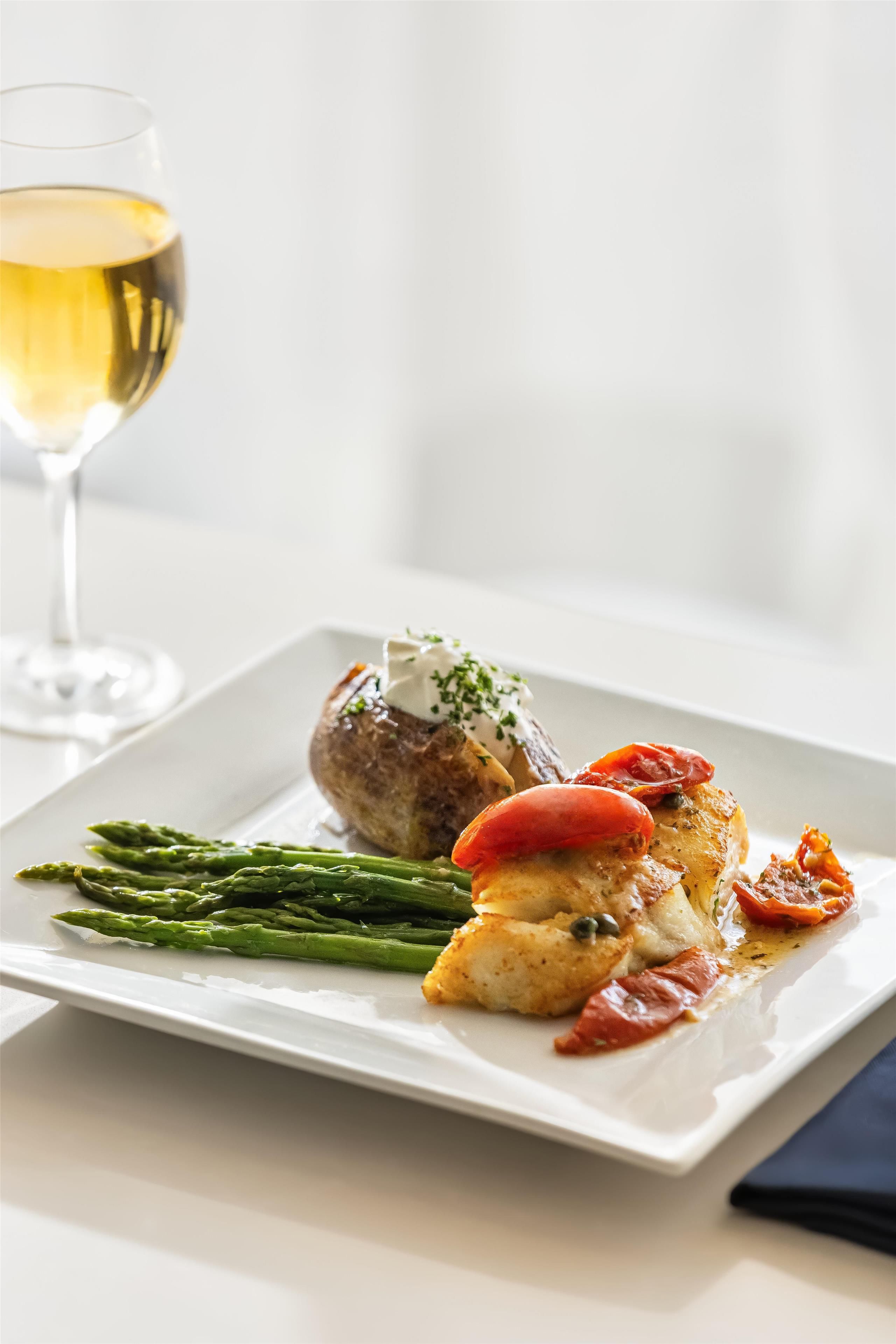 A white plate with a roasted chicken, asparagus, and a red tomato on a white table.