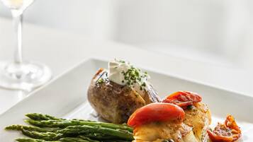 A white plate with a roasted chicken, asparagus, and a red tomato on a white table.