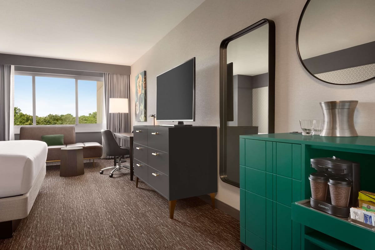 A hotel room with a bed, a desk, a chair, a TV, a dresser, a mirror, and a view of the outdoors.
