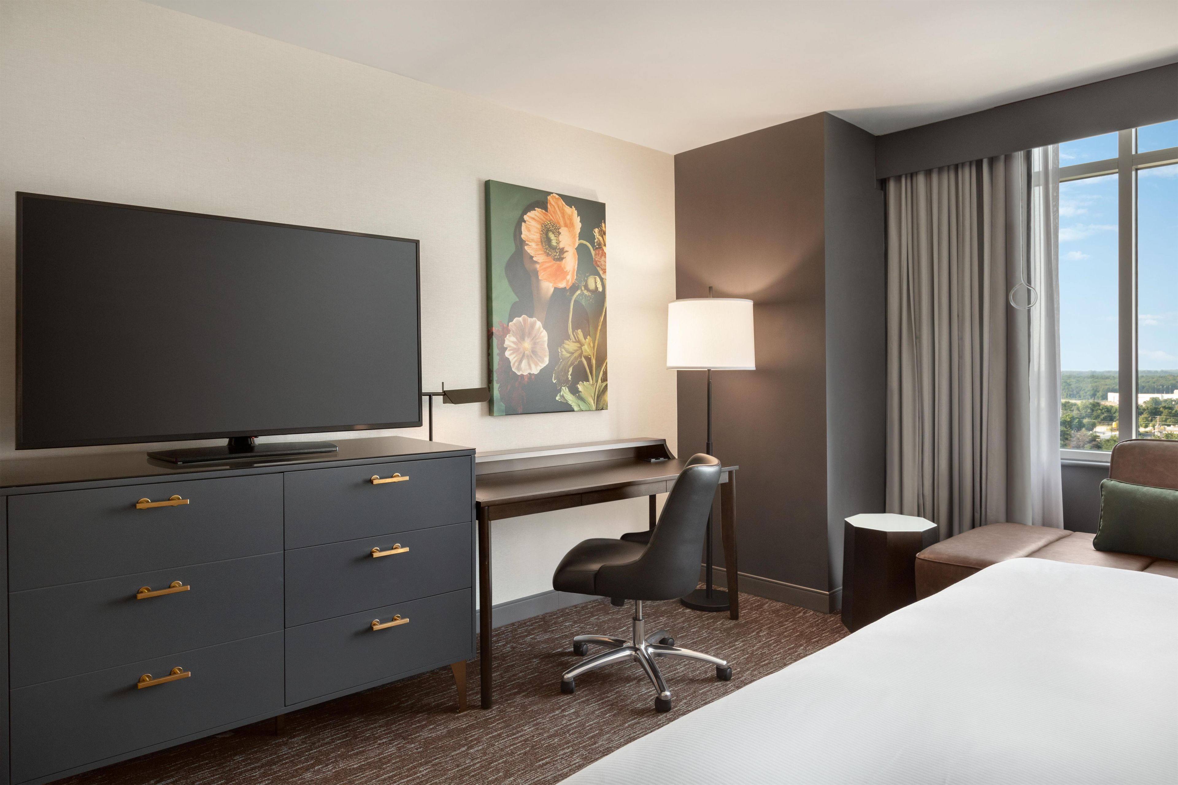 Habitación de hotel con televisión de pantalla plana, escritorio, silla, cama, lámpara y un cuadro en la pared.