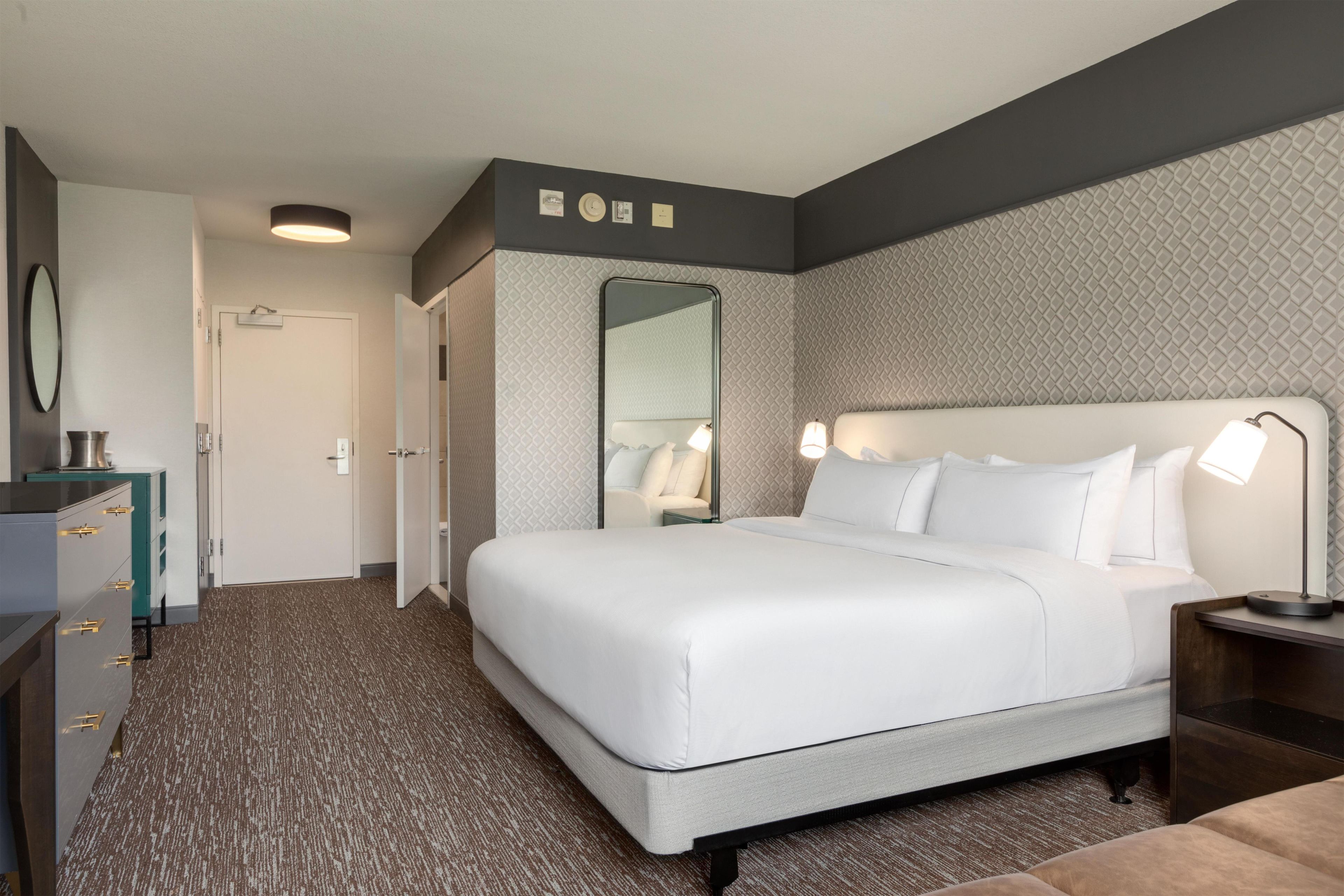 A hotel room with a large bed, a bedside table with a lamp, a dresser, and a mirror on the wall.