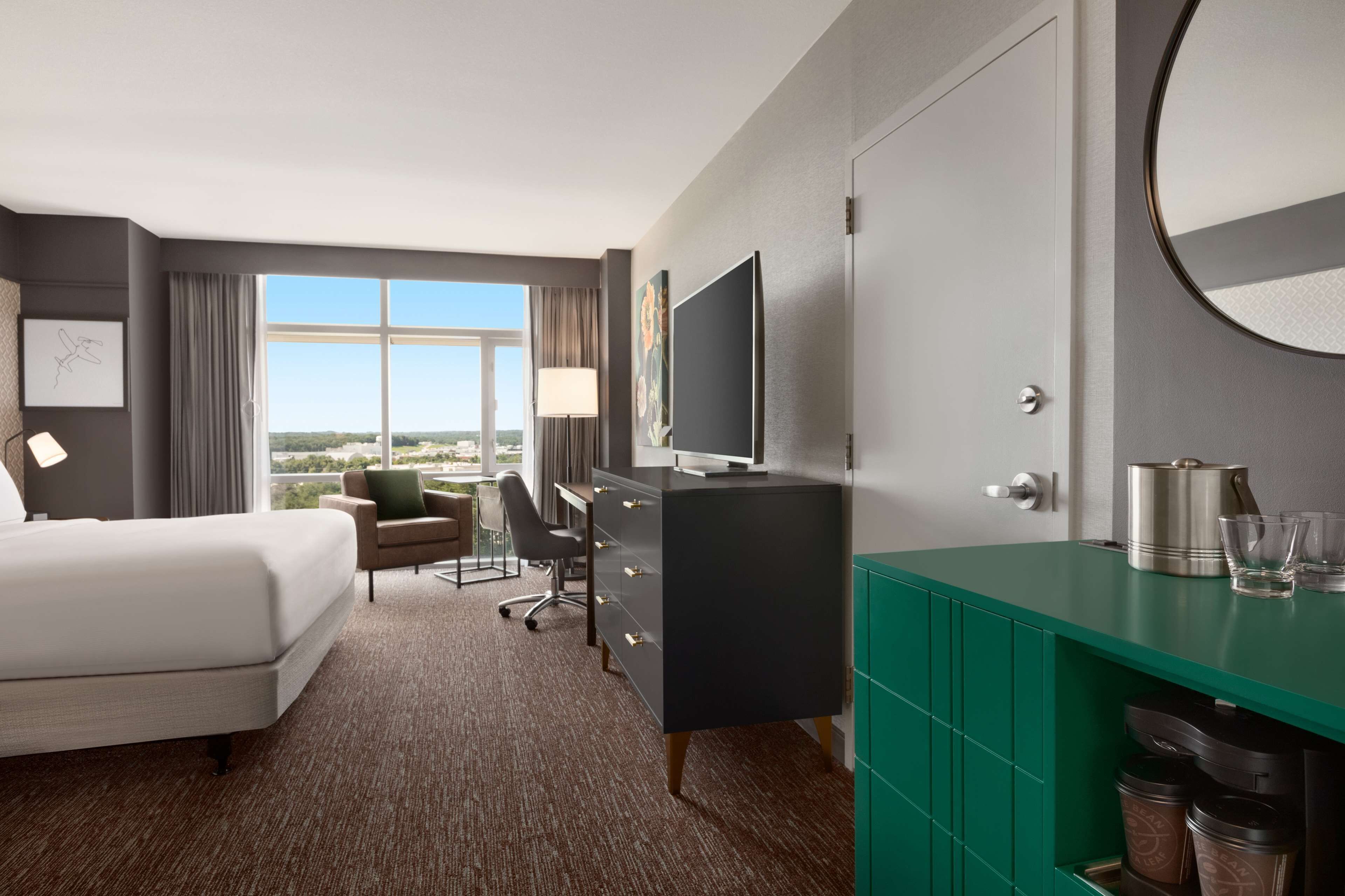 A hotel room with a bed, desk, chair, TV, and a view of the city.