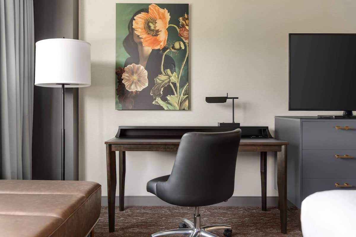 A hotel room with a desk, a chair, a flat-screen TV, a lamp, and a painting of flowers on the wall.