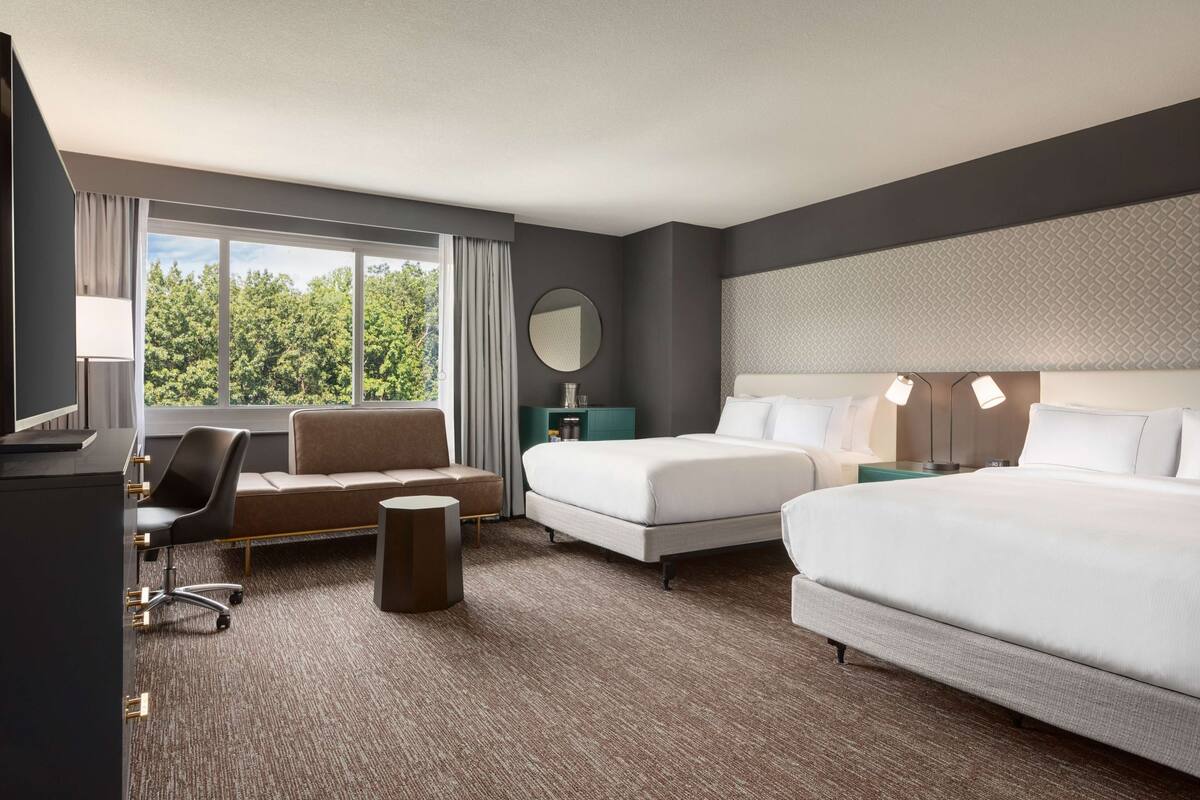 A hotel room with a bed, a desk, a chair, a round mirror, and a view of trees through a window.