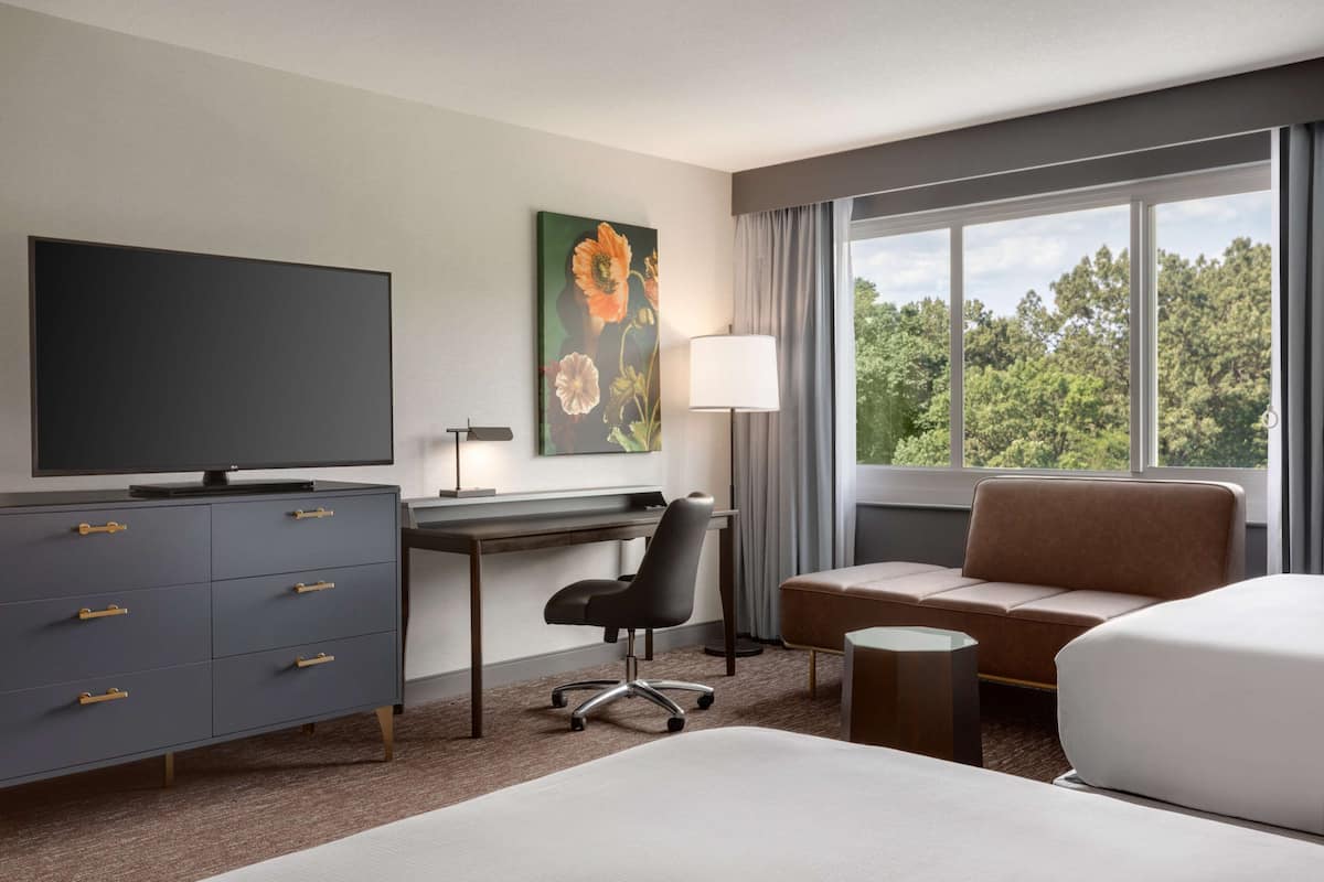 A hotel room with a bed, a desk, a chair, a TV, a painting, and a window with a view of trees.