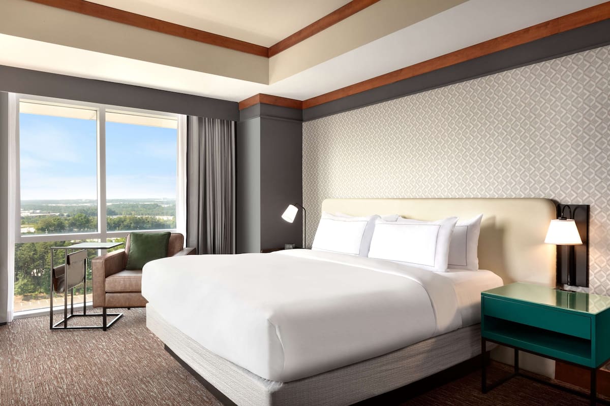 A hotel room with a large bed, a nightstand with a lamp, a chair, and a view of the outdoors.