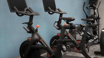 Stationary bikes with digital screens and the Peloton logo, arranged in a gym with a blue wall and exercise equipment in the background.
