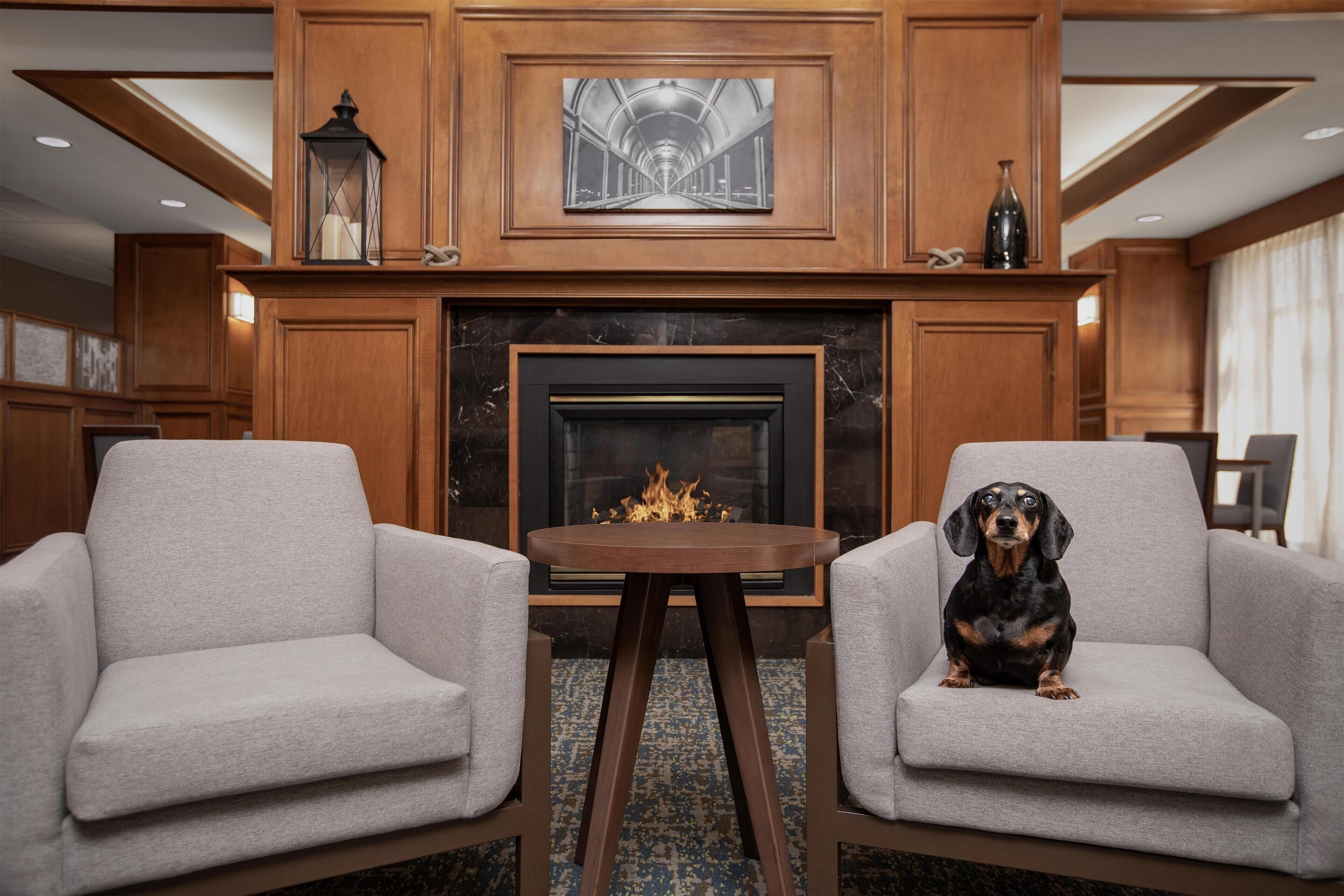 A room with two grey armchairs, a small table, a fireplace, and a framed artwork above the fireplace.