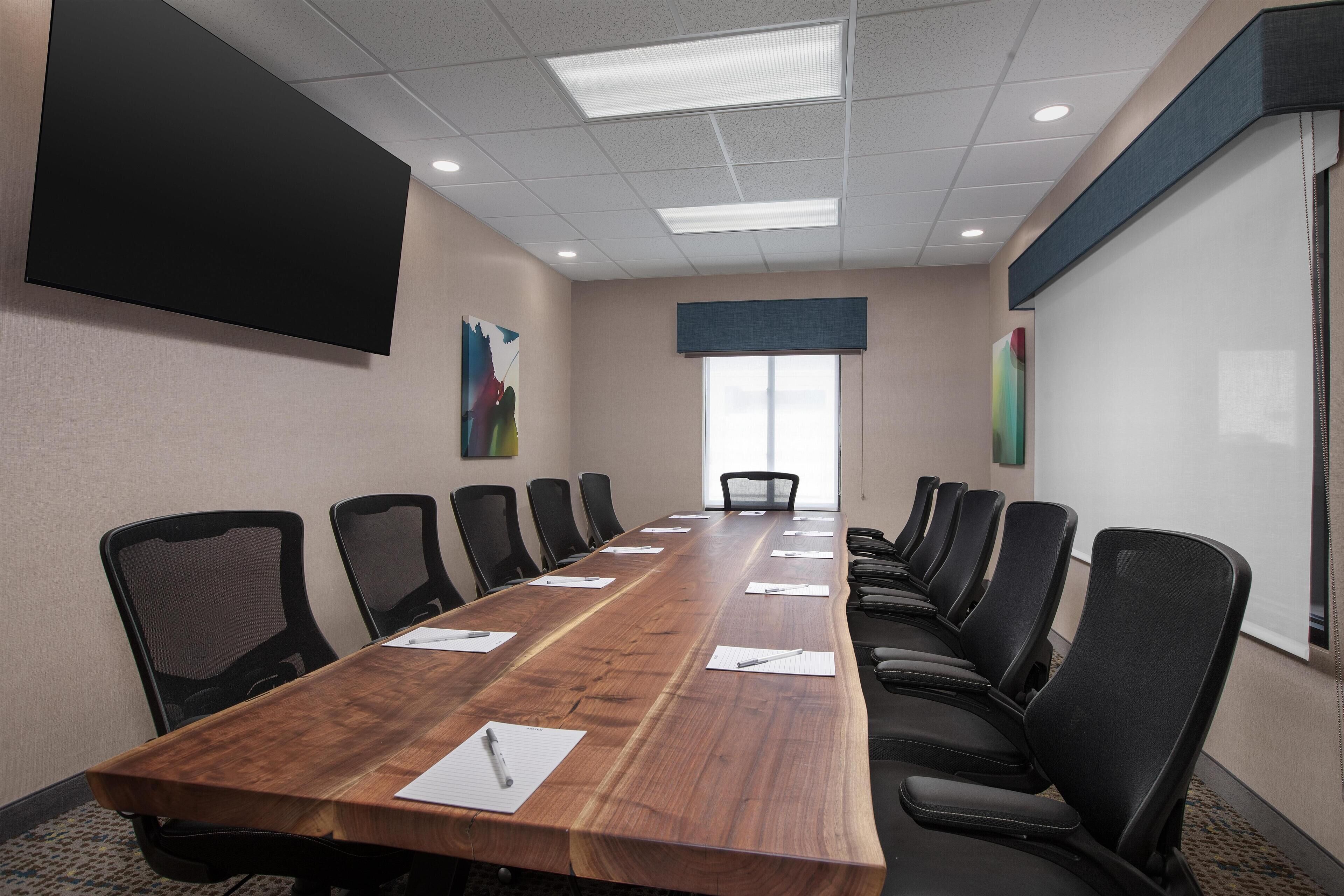 Un salón de conferencias con una larga mesa de madera, sillas negras, una pantalla grande y un ventanal con persianas.
