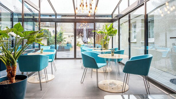 Een moderne, overdekte patio met glazen wanden, blauwe stoelen en potplanten.