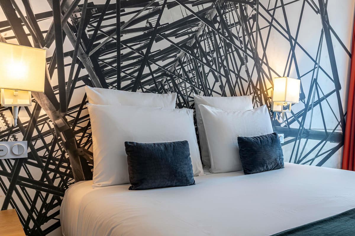 Una habitació d'hotel amb un disseny modern, que destaca un llit amb roba de llit blanca i almoades fosques, una tauleta de nit de fusta i un dibuix geomètric a la paret.