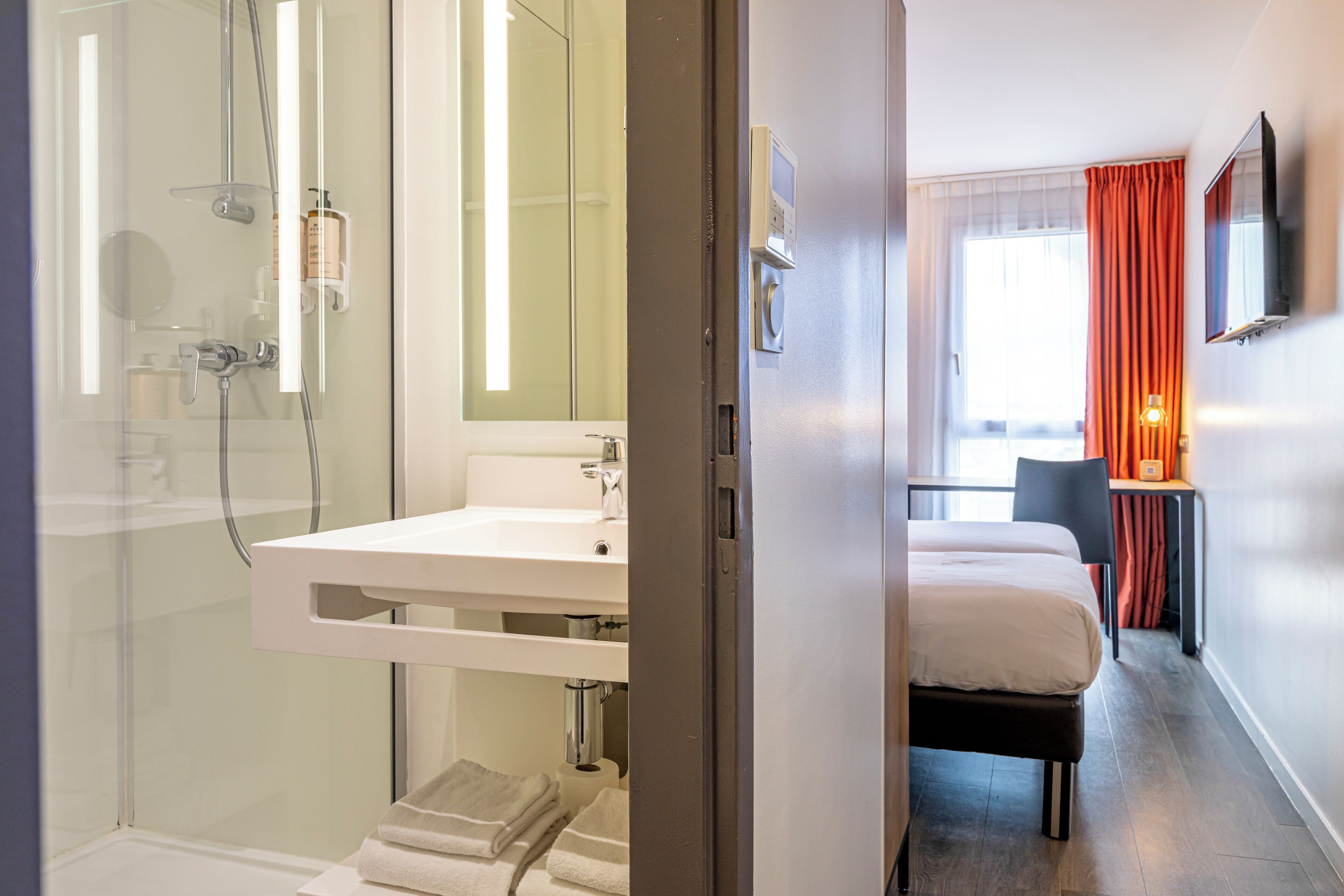 Hotelkamer met een douche in een glazen cabine, een witte wastafel, een bed, een bureau met een stoel en een aan de muur bevestigde televisie.