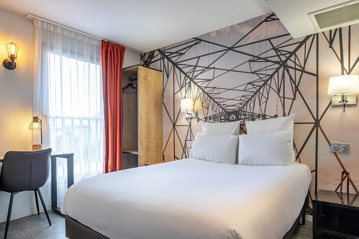 Una habitació d'hotel amb un llit gran, un escriptori i una cadira. Hi ha una finestra amb cortines, una paret amb un disseny geomètric i una làmpada penjada a la paret.