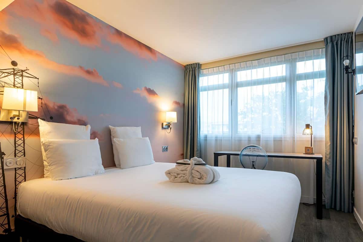 Habitació d'hotel amb un llit, una taula de treball, una làmpada i un mural a la paret.