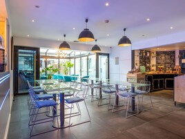 Un comedor moderno con mesas de vidrio y sillas de metal, iluminación colgante y vistas al exterior a través de grandes ventanales.