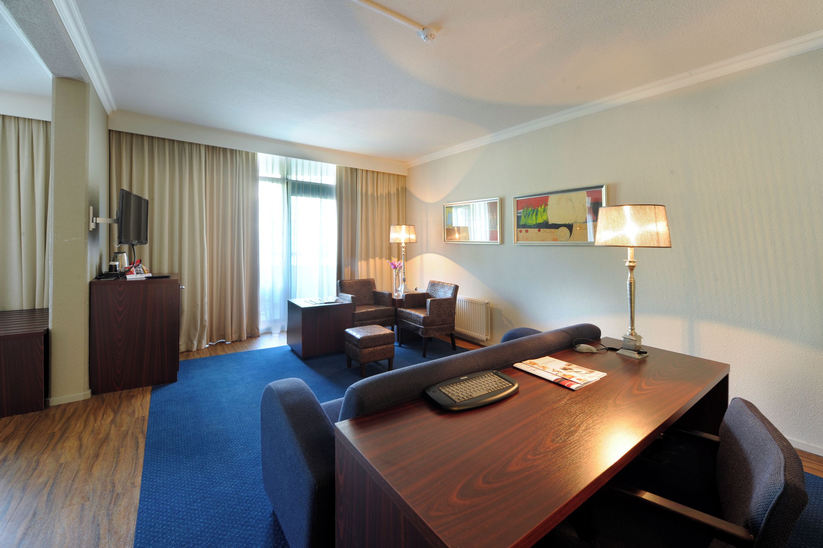 Habitación de hotel con escritorio de madera, dos sillas, un sofá, un televisor, una lámpara y una ventana con cortinas.