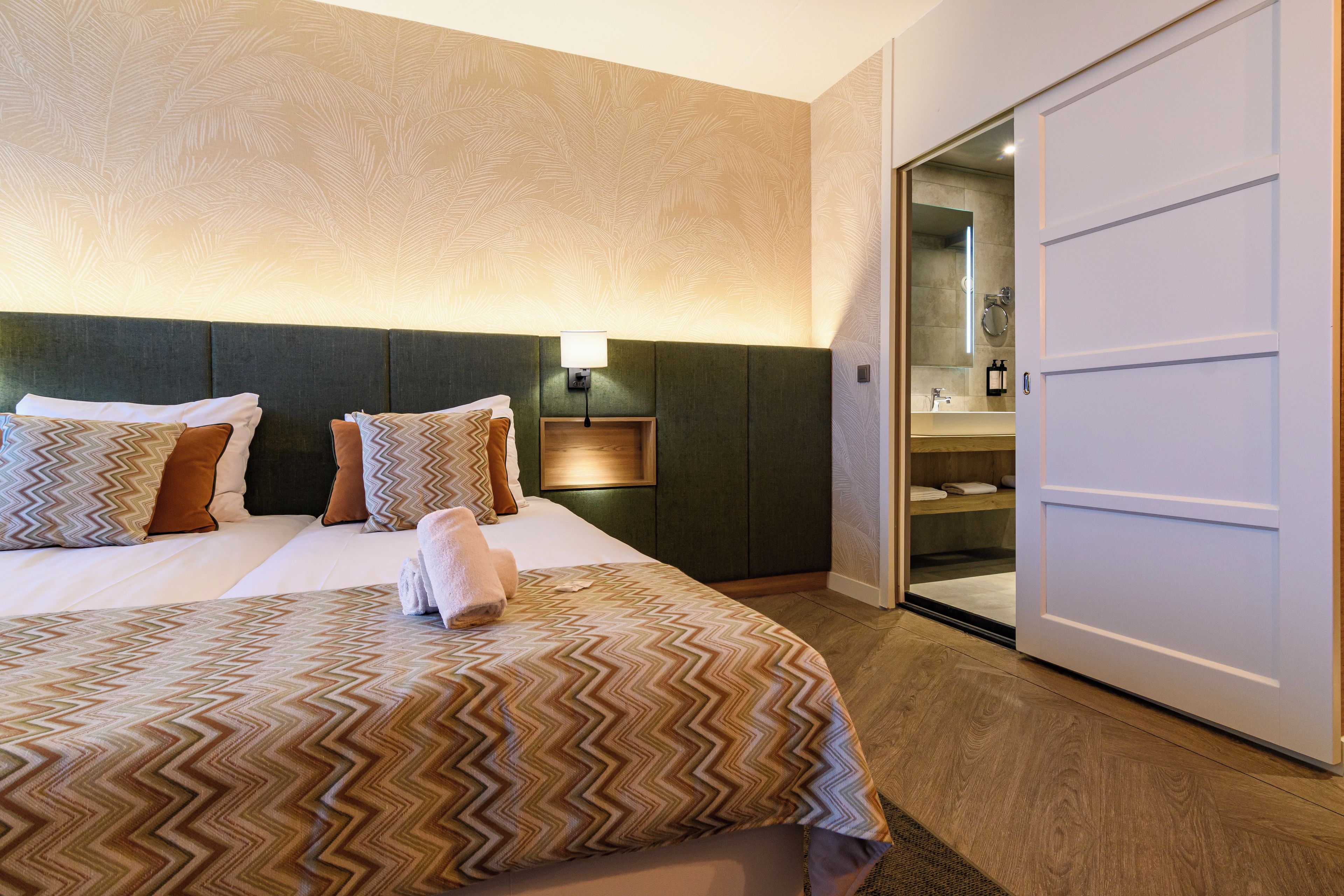 A hotel room with a bed, pillows, a nightstand, a lamp, and a bathroom visible through an open door.