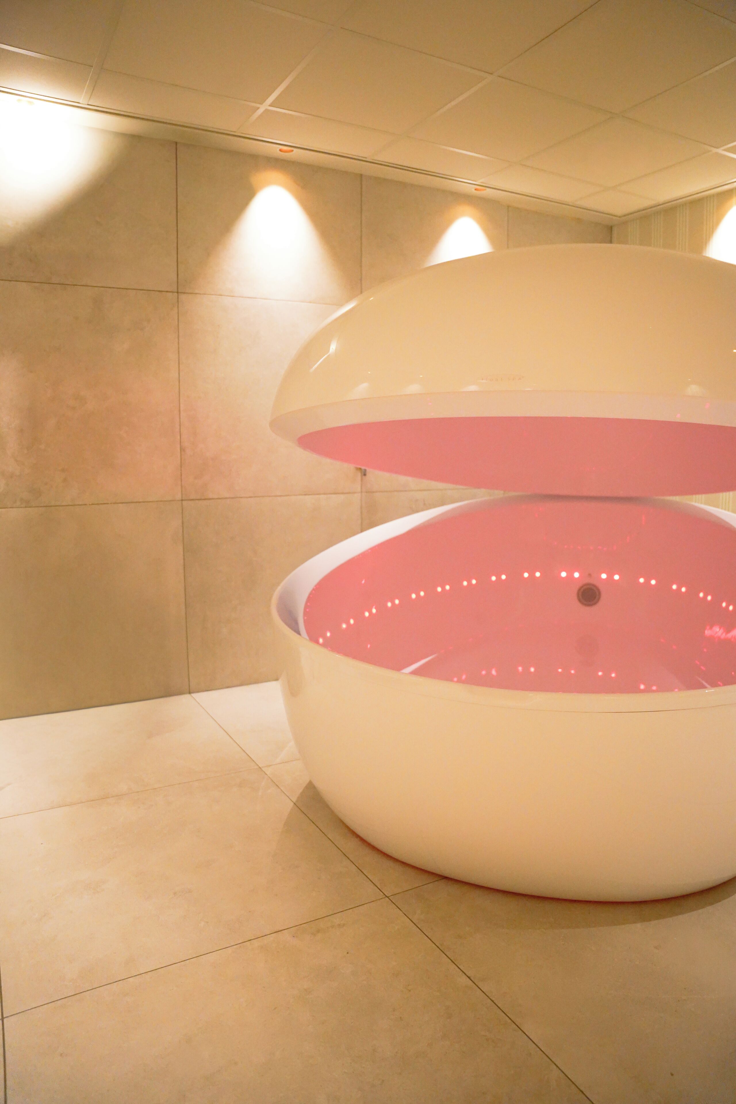 A large, white, circular structure with a pink interior, possibly a water feature, in a tiled room with ceiling lights.