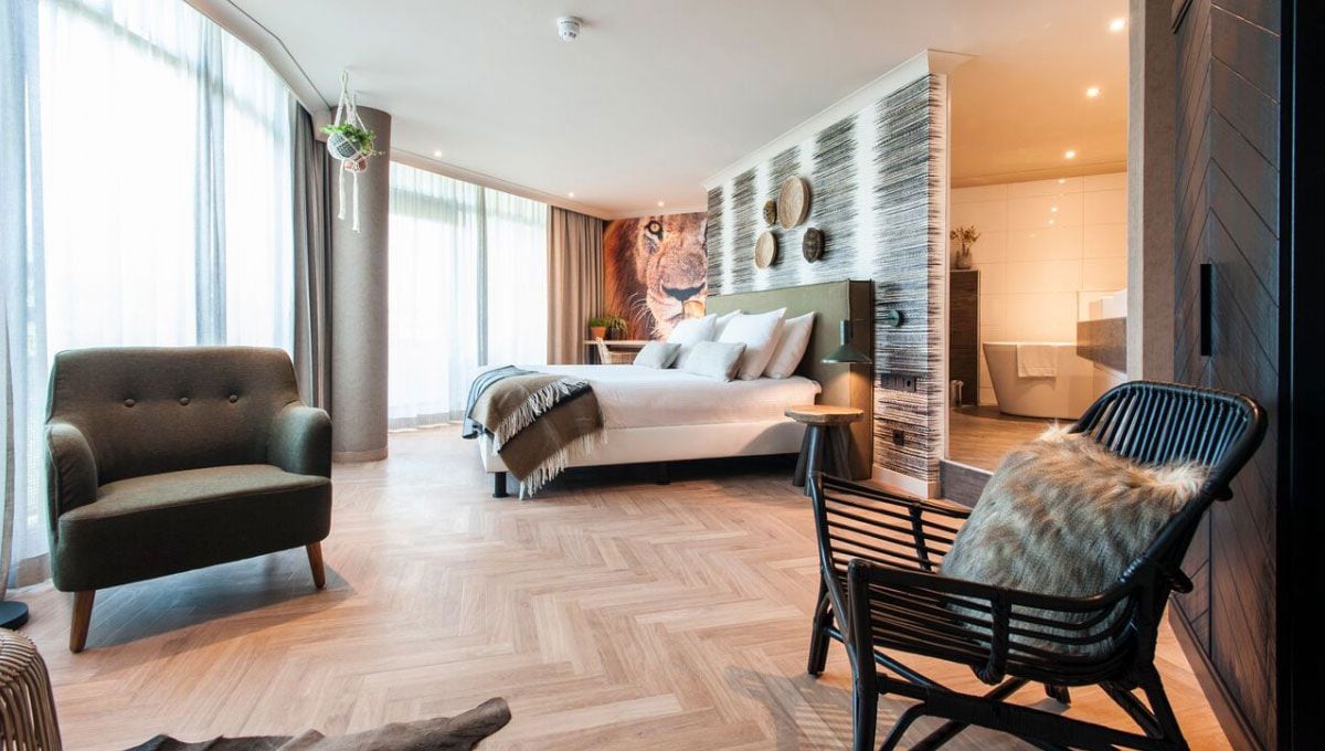 Habitación de hotel moderna con una cama grande, una zona de estar con una silla, una mesita y vistas al baño.