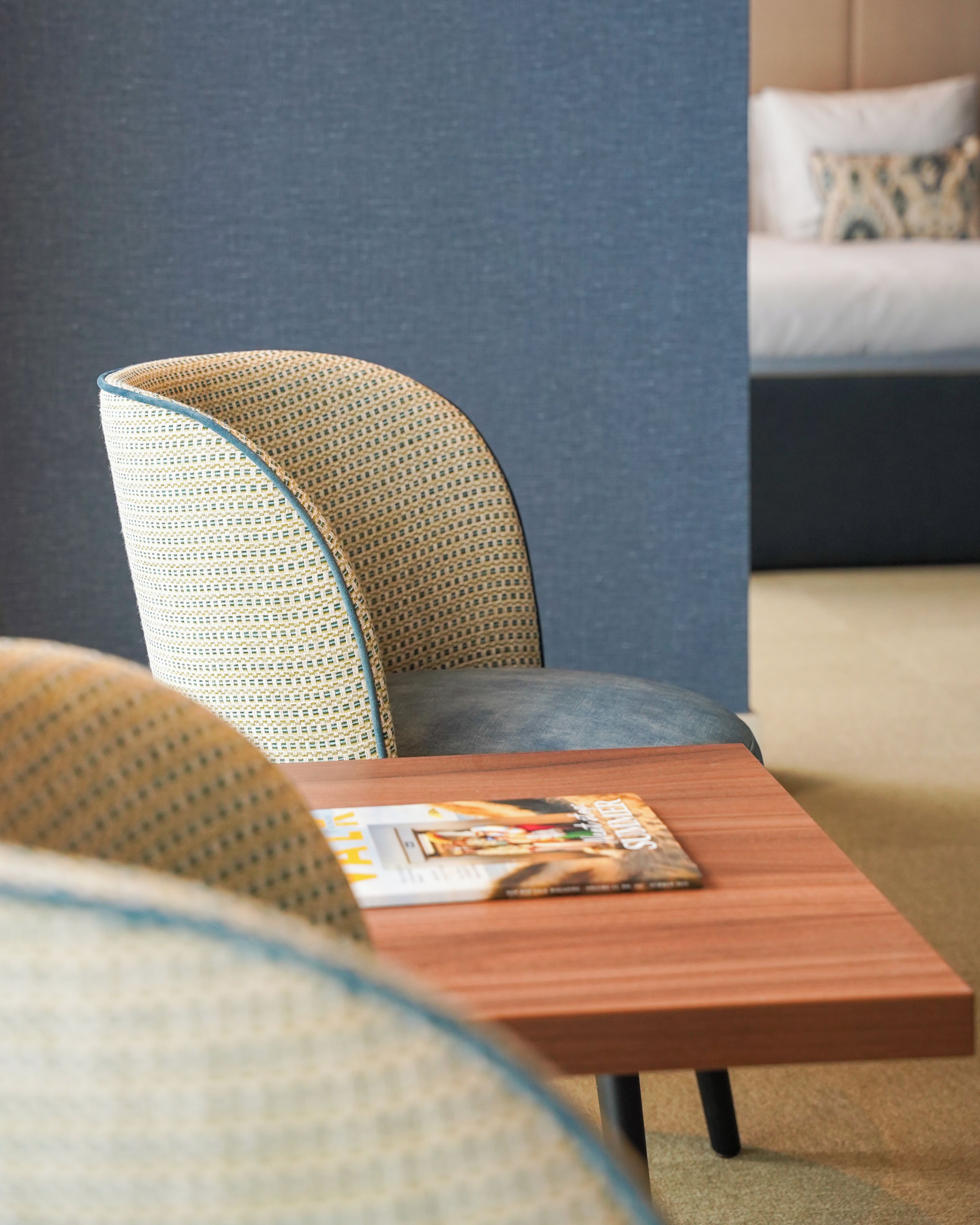 Una habitación moderna con una butaca curvada tapizada, una mesita de madera con una revista y una cama con un cojín estampado al fondo.