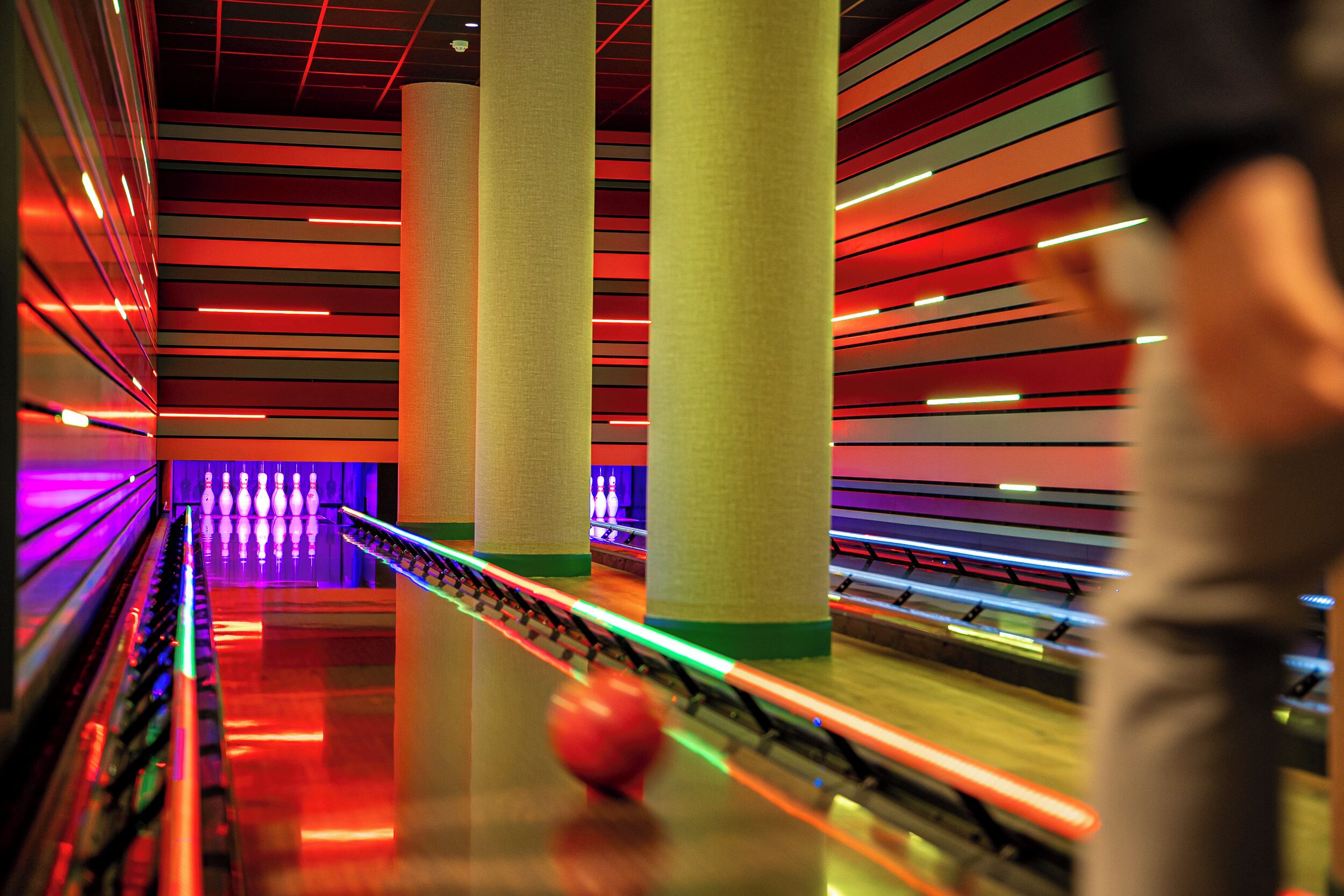 A bowling alley with multiple lanes, a bowling ball in motion, and a person in the foreground.