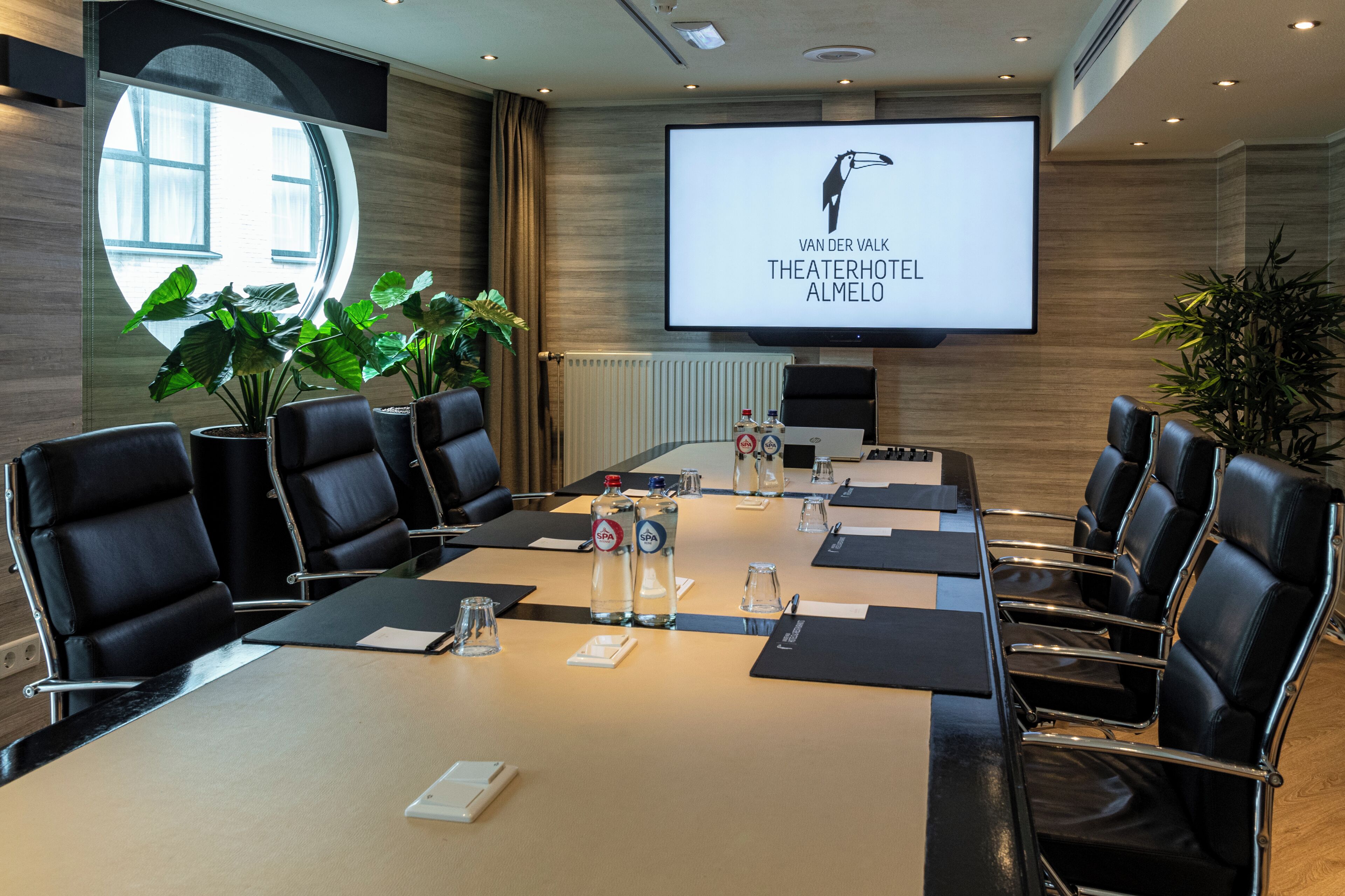Un salón de conferencias con una pantalla grande mostrando 'VAN DER VALK THEATERHOTEL ALMELO'.