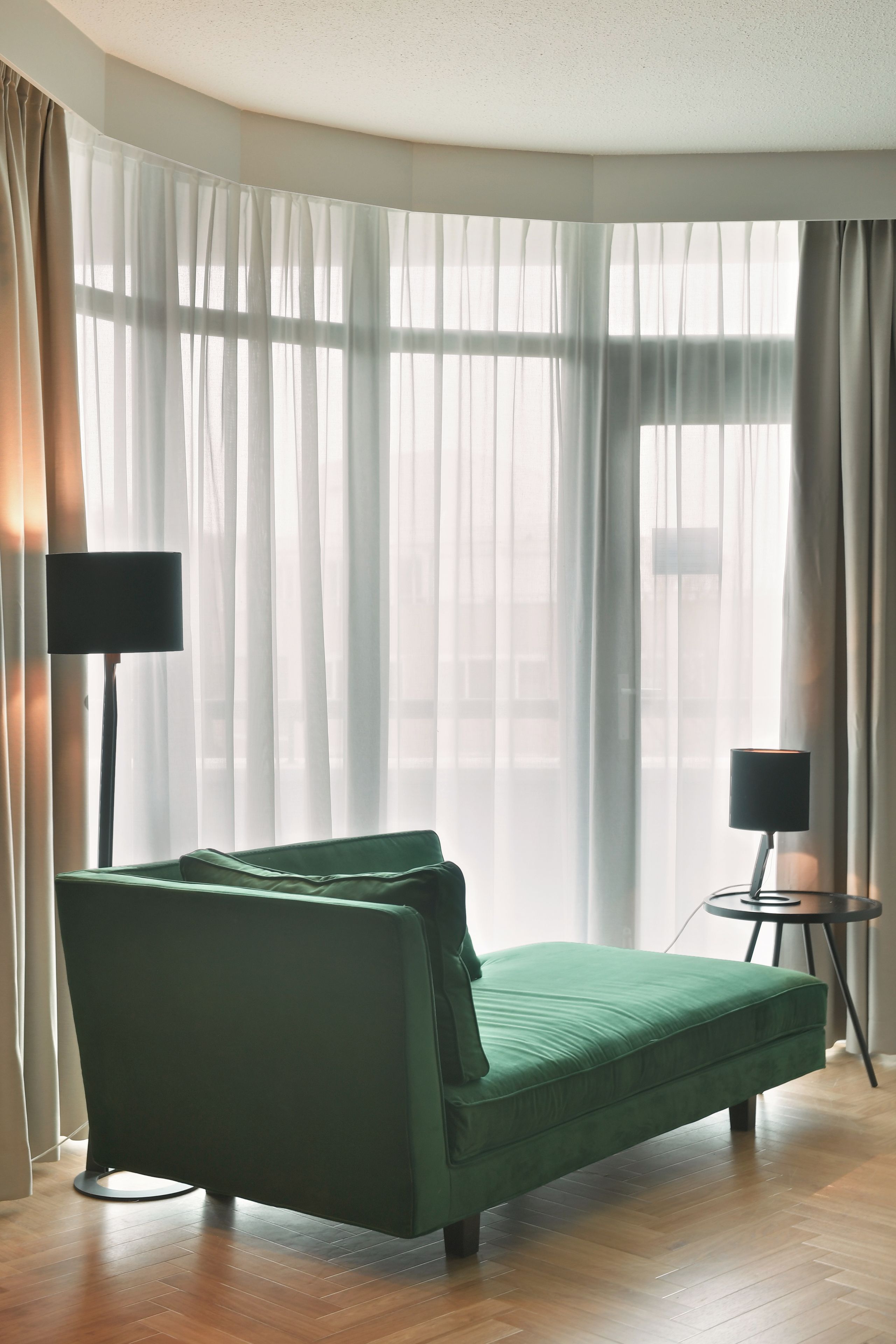 A room with a green sofa, a small side table, a lamp, and sheer curtains.