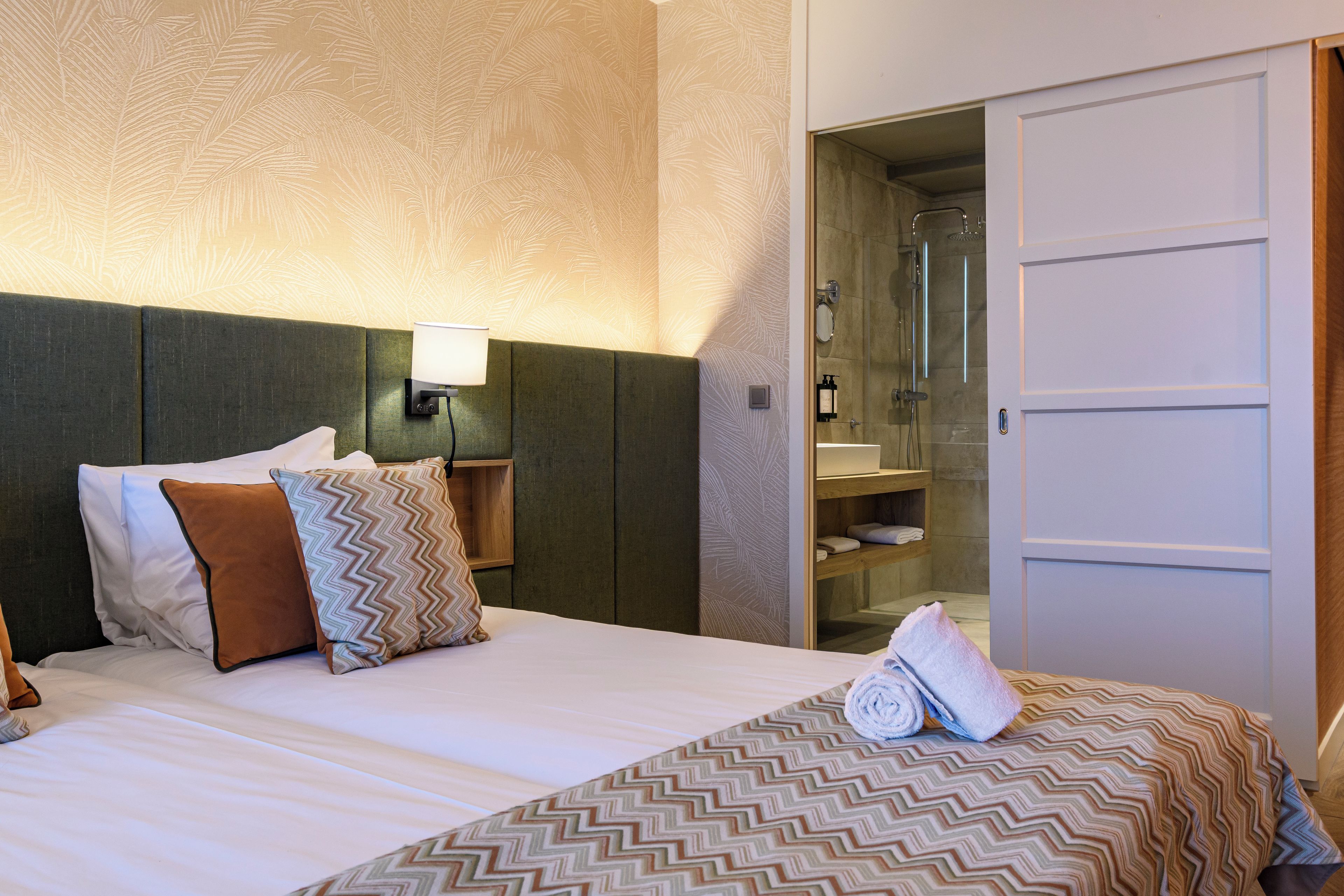 Habitación de hotel con cama, almohadas, mesita de noche, lámpara de pared y puerta abierta que da a un baño con ducha y espejo.