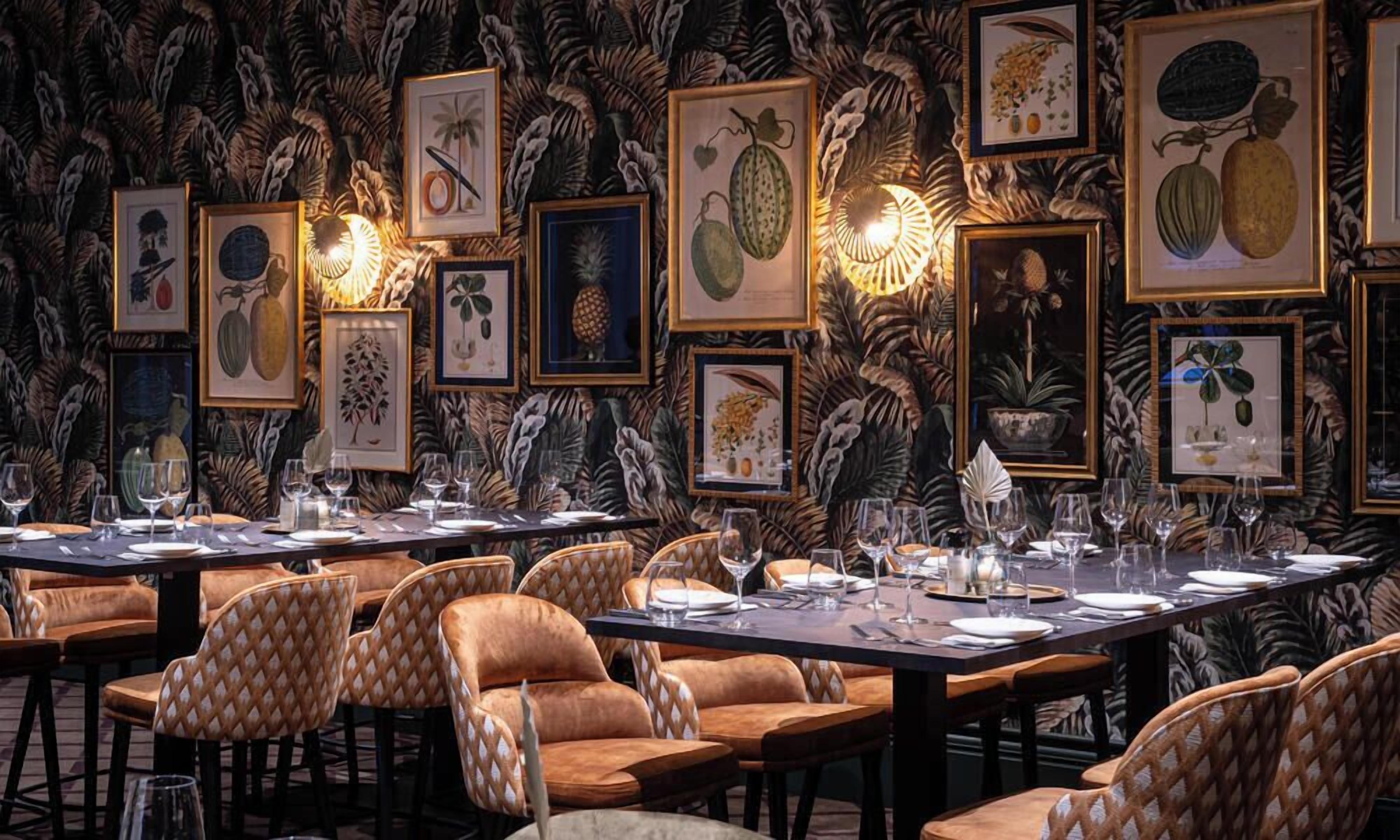 Een restaurant met een lange tafel gedekt voor een maaltijd, met botanische illustraties in de wand en comfortabele stoelen.