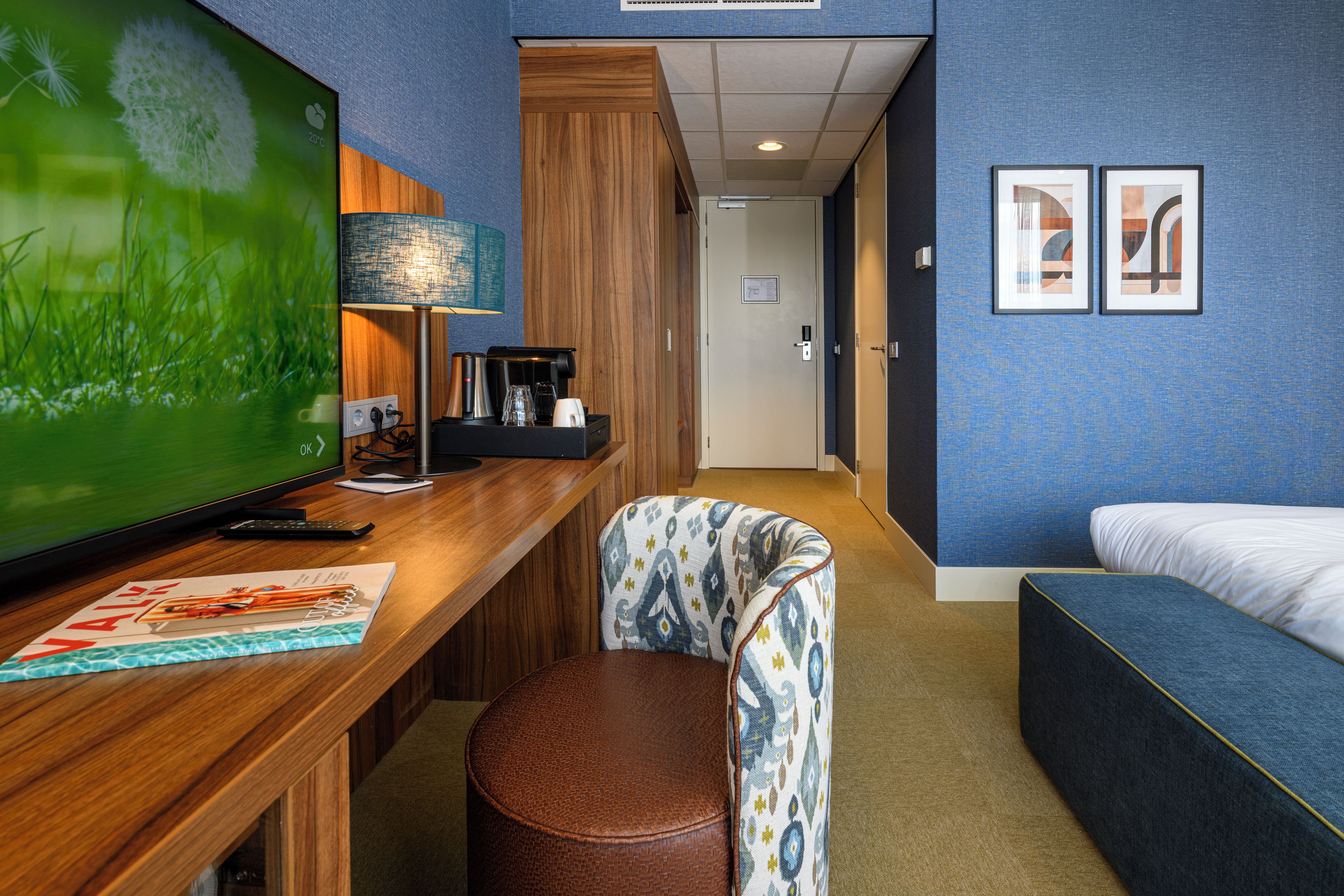 Habitación de hotel con un escritorio de madera, un televisor de pantalla plana grande, una cafetera, una silla estampada, una cama y dos cuadros en la pared.