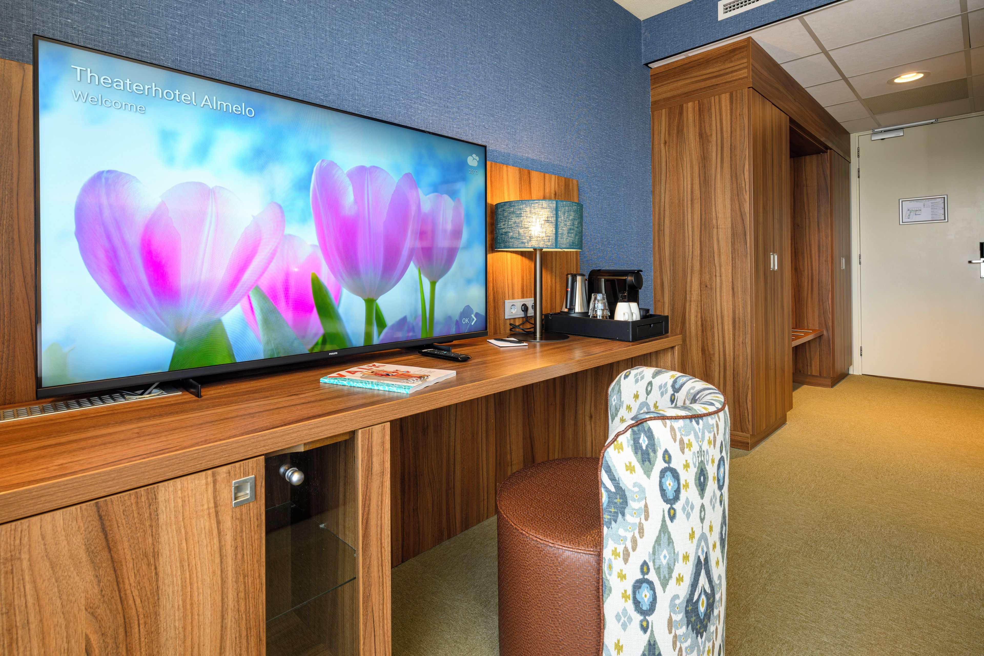 Un espace d'accueil d'hôtel avec un téléviseur diffusant une image florale, une machine à café et un coin salon agrémenté d'un pouf à motifs.