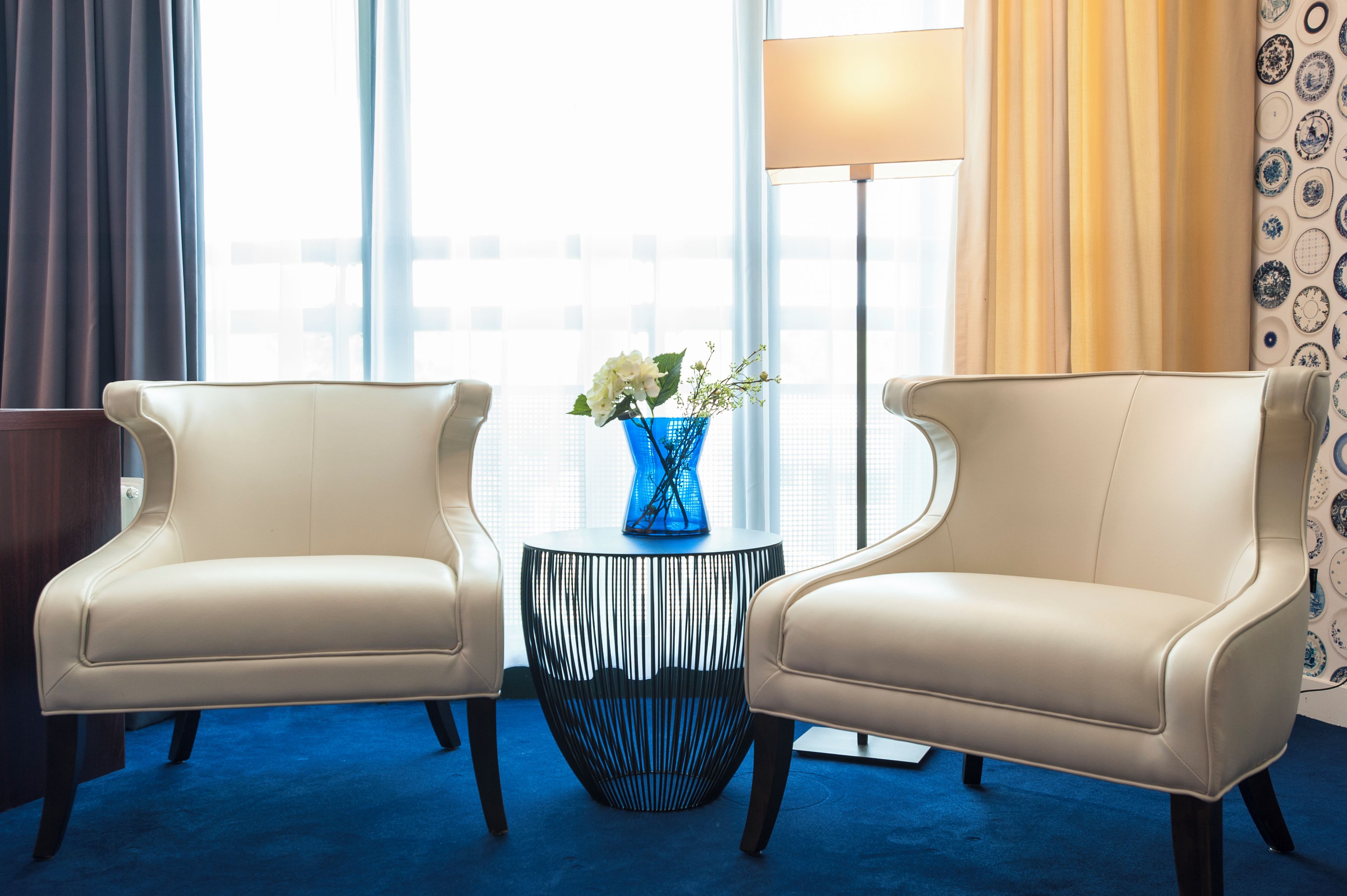 A hotel room with two armchairs, a side table with a vase of flowers, a floor lamp, and a view of the city through large windows.