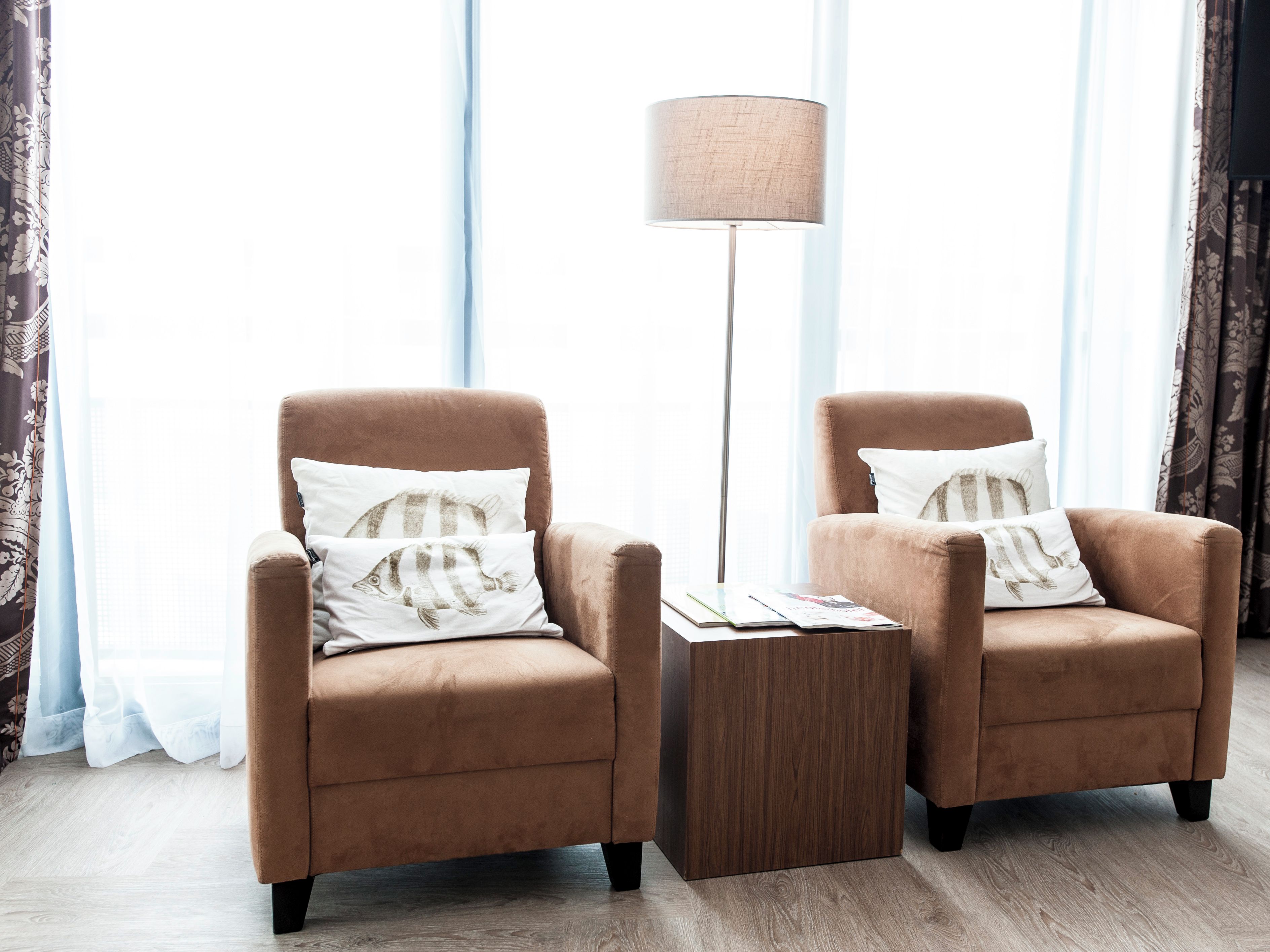 Two brown armchairs with striped pillows, a wooden side table, and a floor lamp in a room with large windows and curtains.