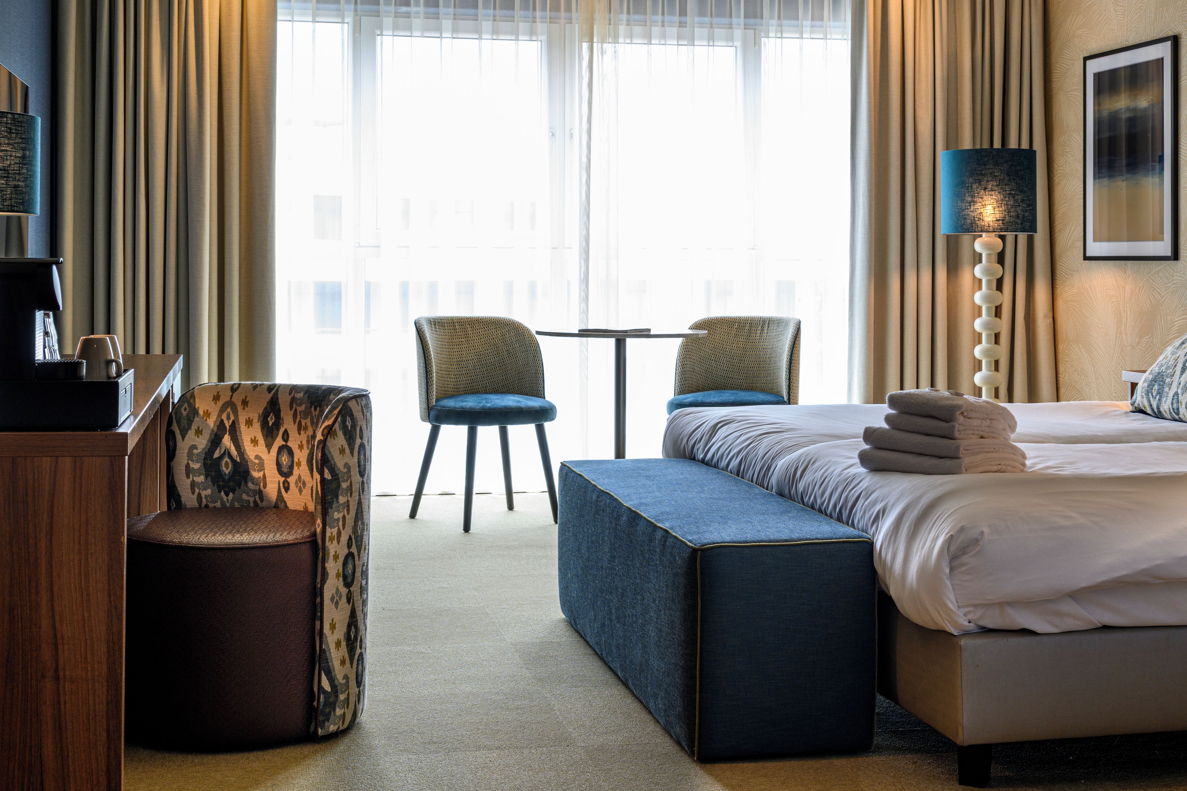 A hotel room with a bed, two chairs, a small table, a coffee machine, and a lamp.