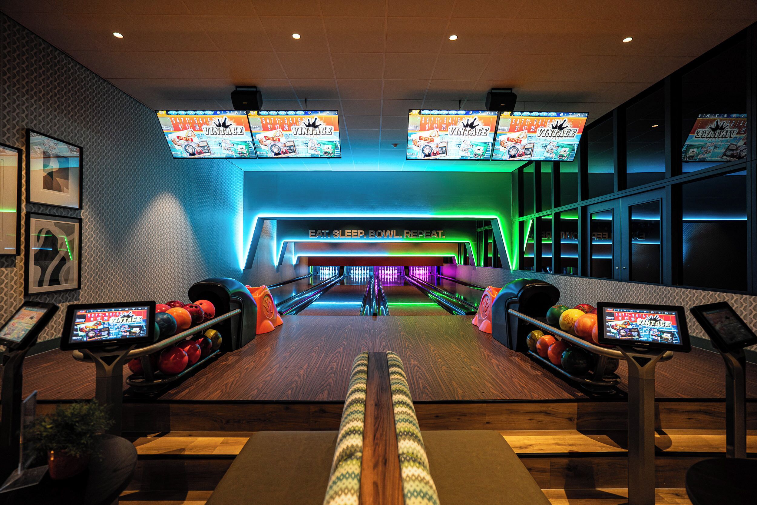 A bowling alley with multiple lanes, overhead screens displaying games, and bowling balls arranged on racks.